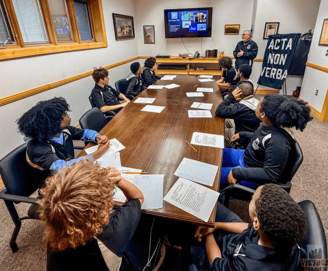 Group of students seated around a long conference table in a classroom, listening to a teacher at the front.