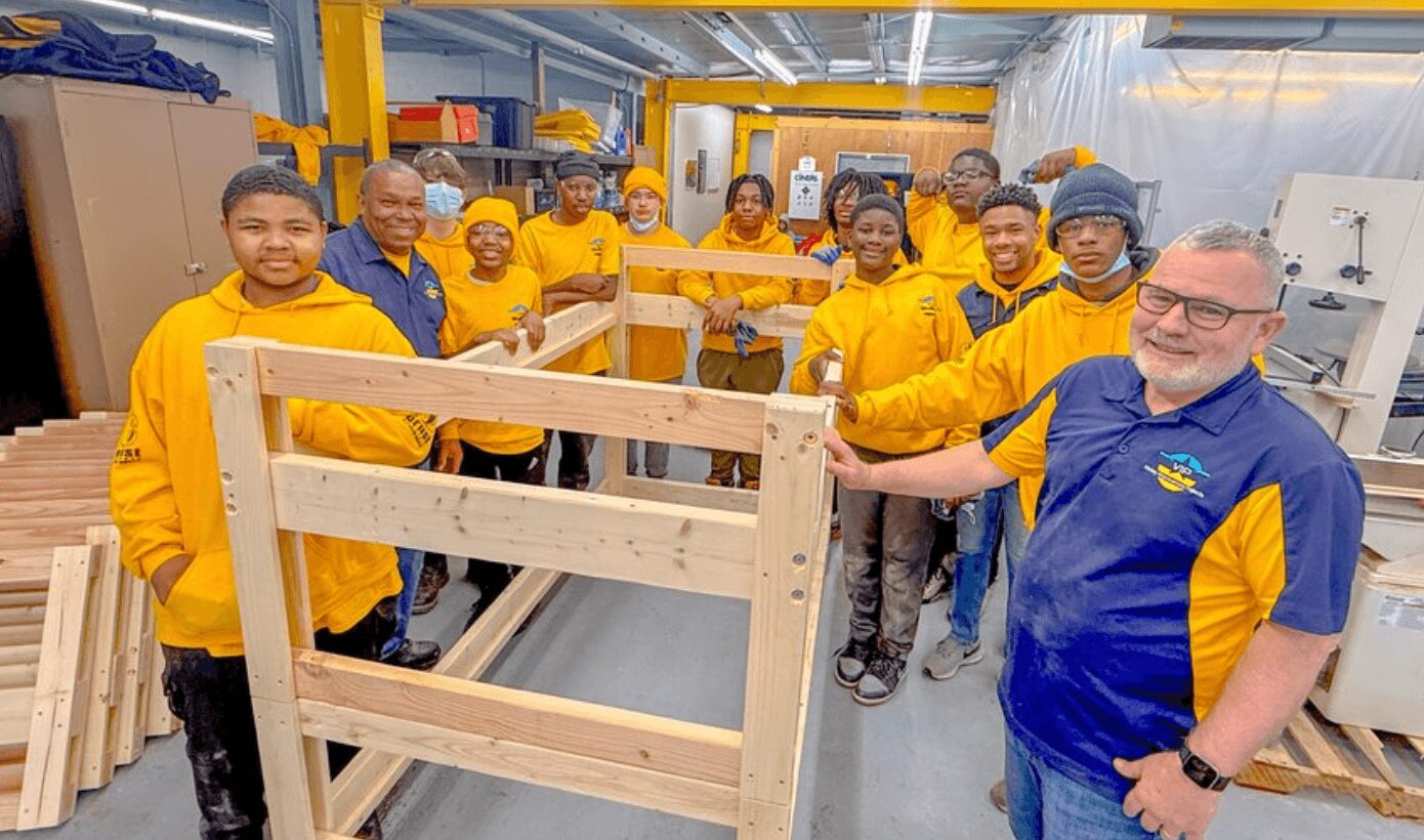 Group of people in yellow and blue uniforms standing in a workshop with wooden furniture pieces.