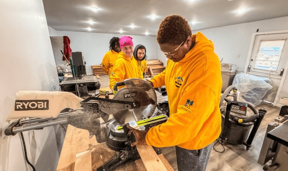 Group of people working on a woodworking project in a workshop, wearing yellow hoodies, with tools and wood around.