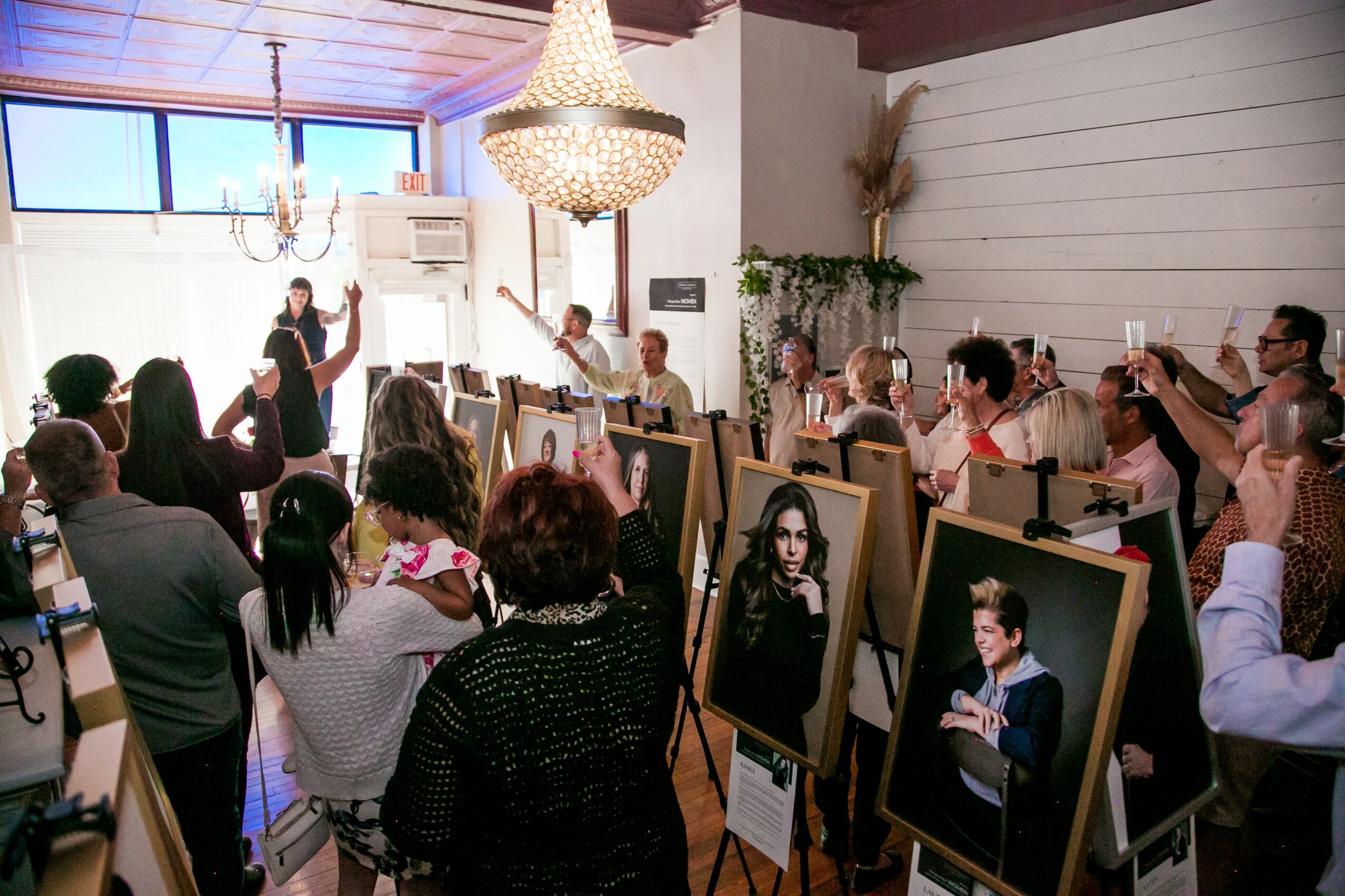 People gathered in a room with framed portraits on easels, some taking photos, chandelier hanging from ceiling, large window.