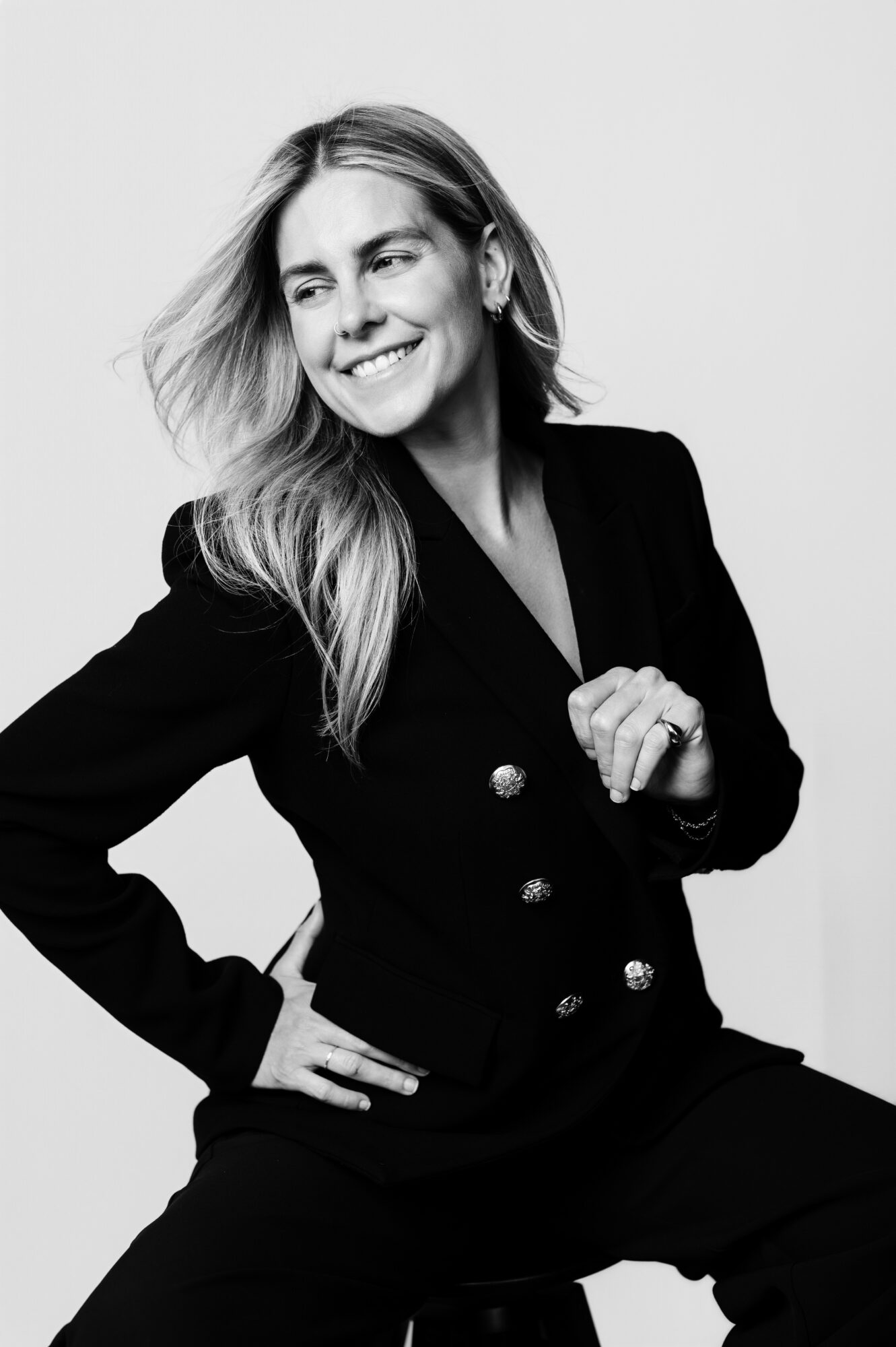 Woman with long hair smiling, wearing a black blazer, sitting with one hand on her hip and the other near her chest.