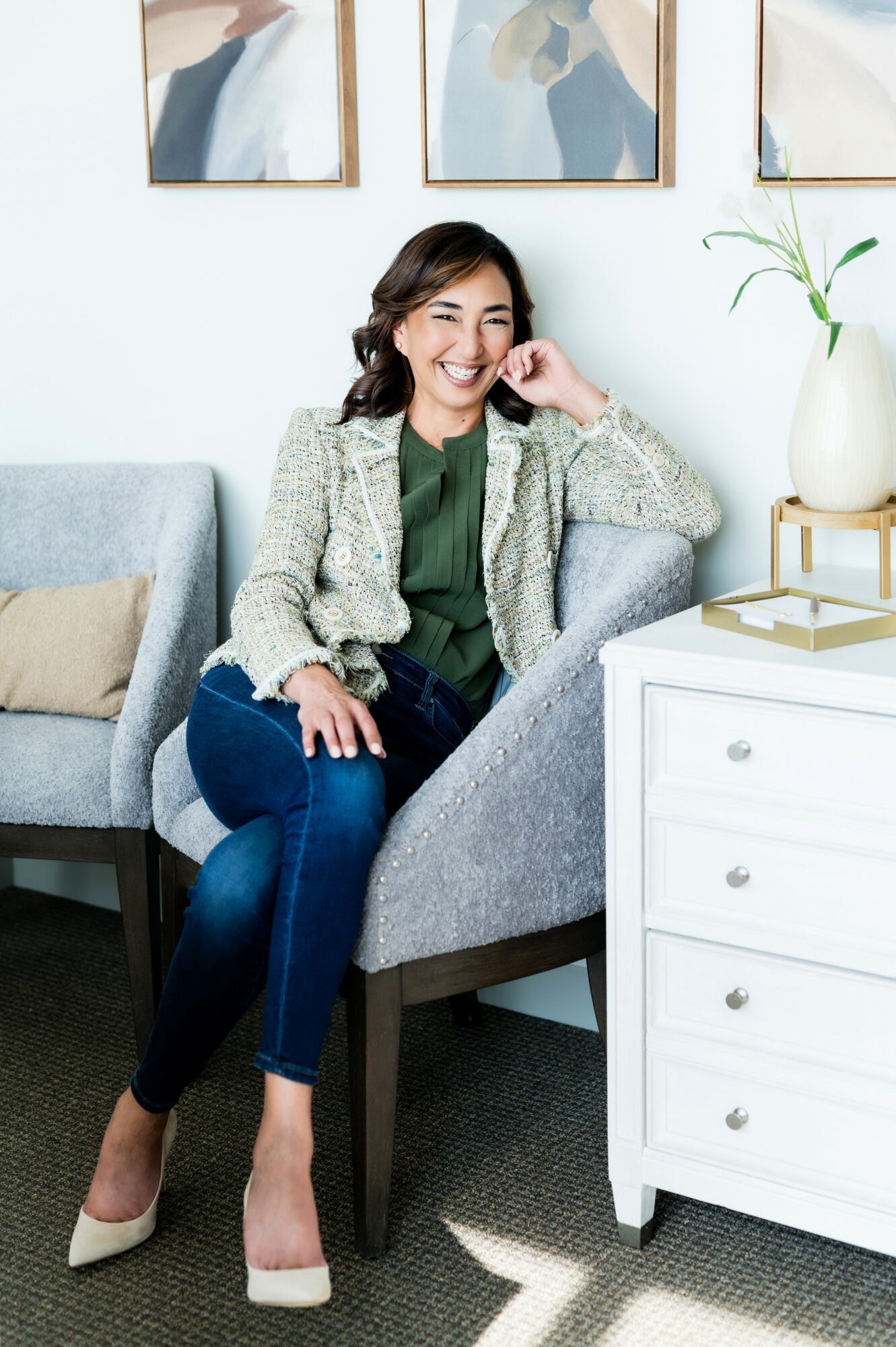 Woman sitting on a gray armchair, smiling, wearing a blazer, green top, and jeans, in a modern room.
