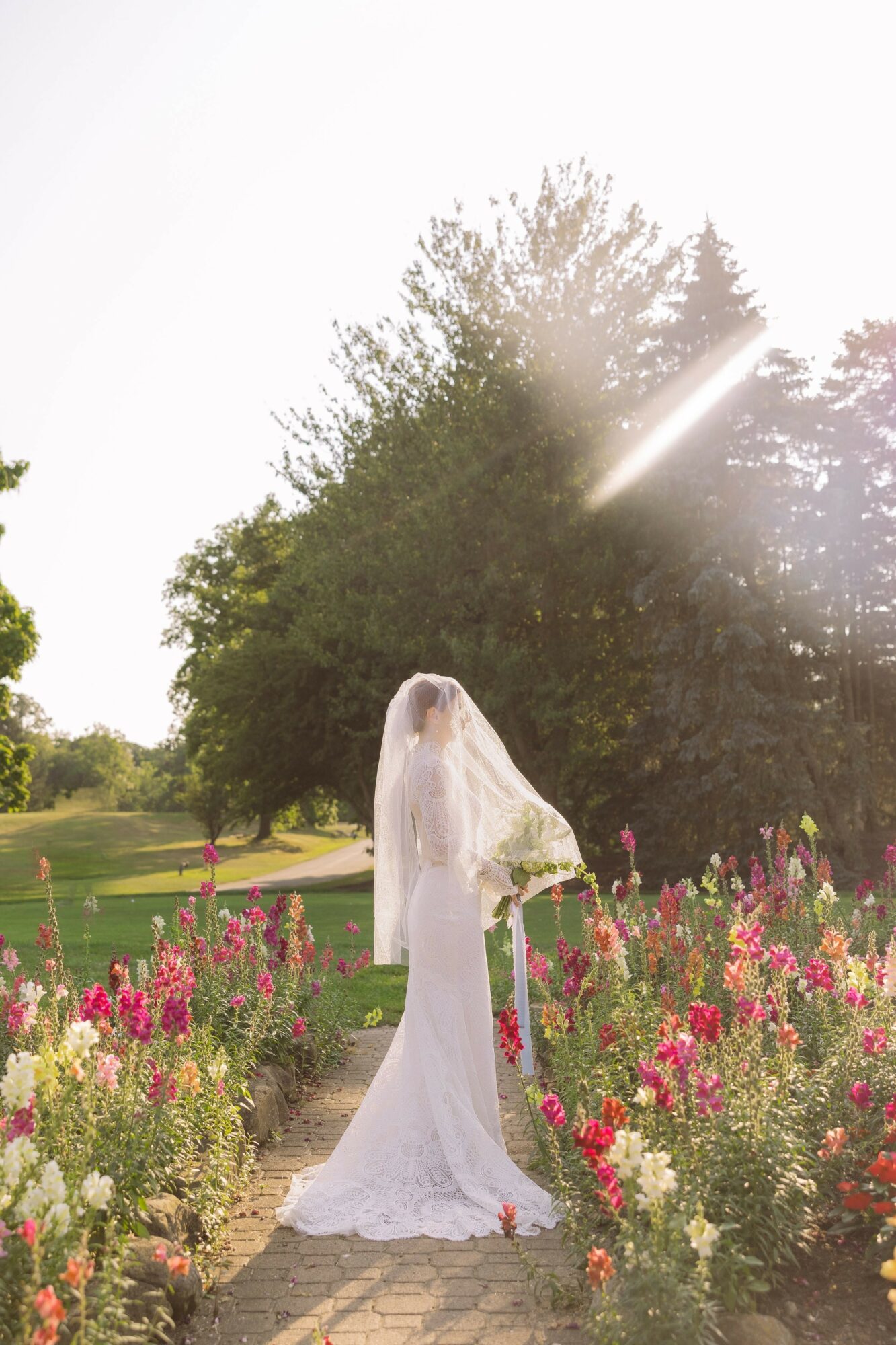 Person in wedding dress and veil standing in a garden with pink and white flowers, trees in the background.