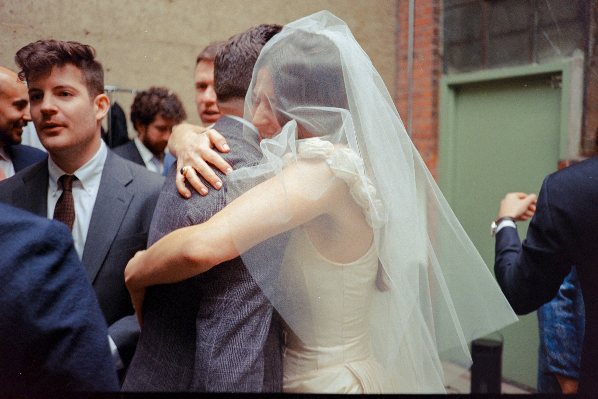 Bride hugging a guest, wearing a white dress and veil, surrounded by people in formal attire outdoors.