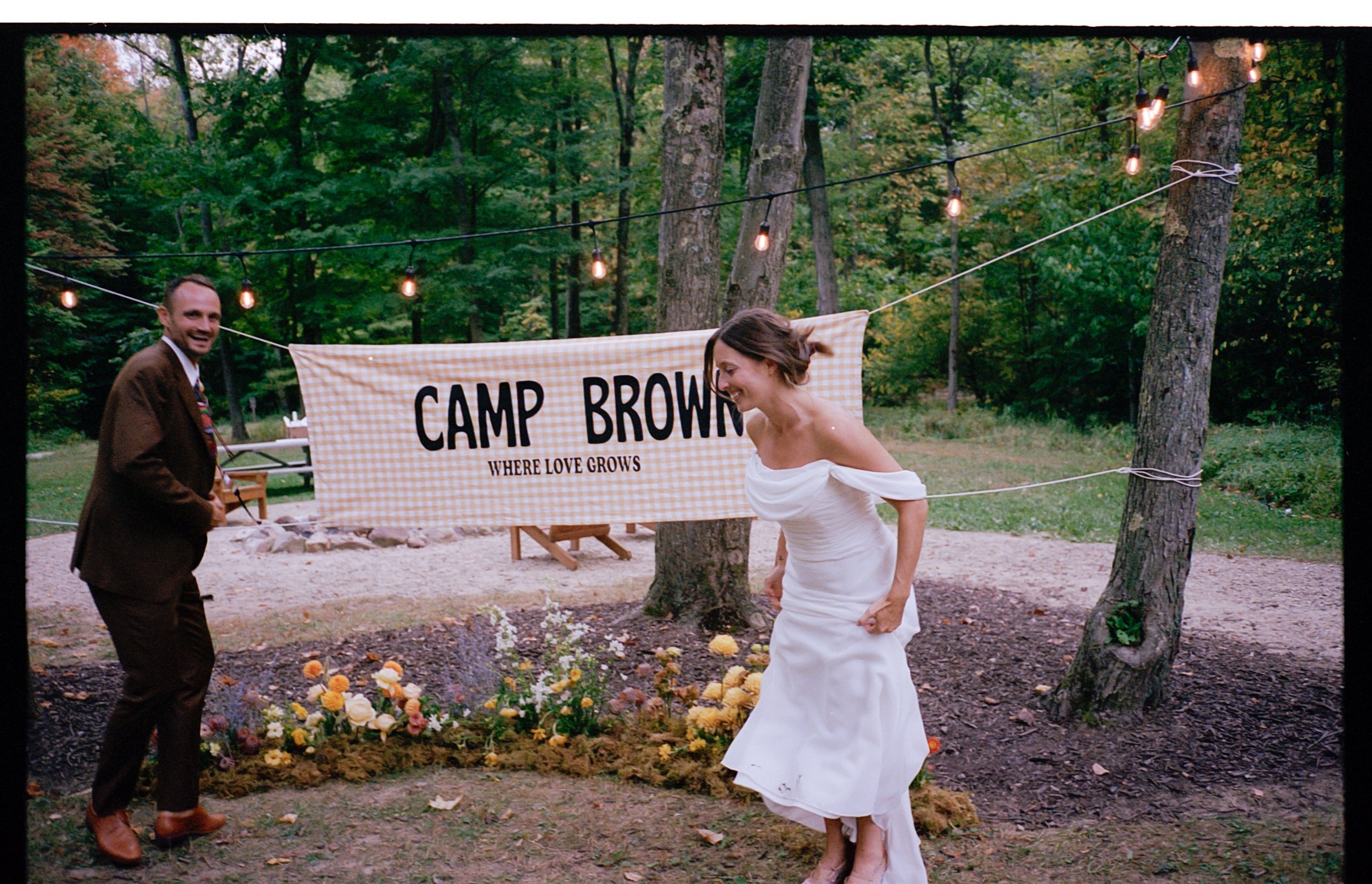 Man and woman holding a banner in a wooded outdoor setting with string lights and flowers.