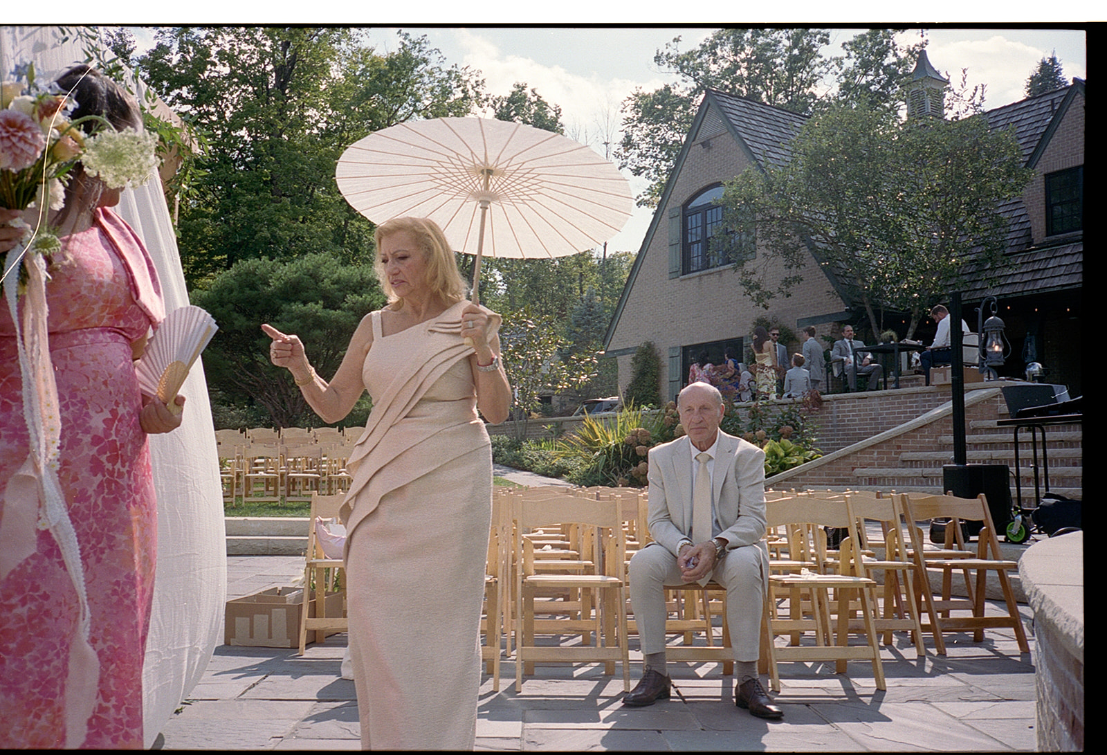 Woman holding a microphone outdoors with a man sitting on chairs nearby, trees and a house in background.
