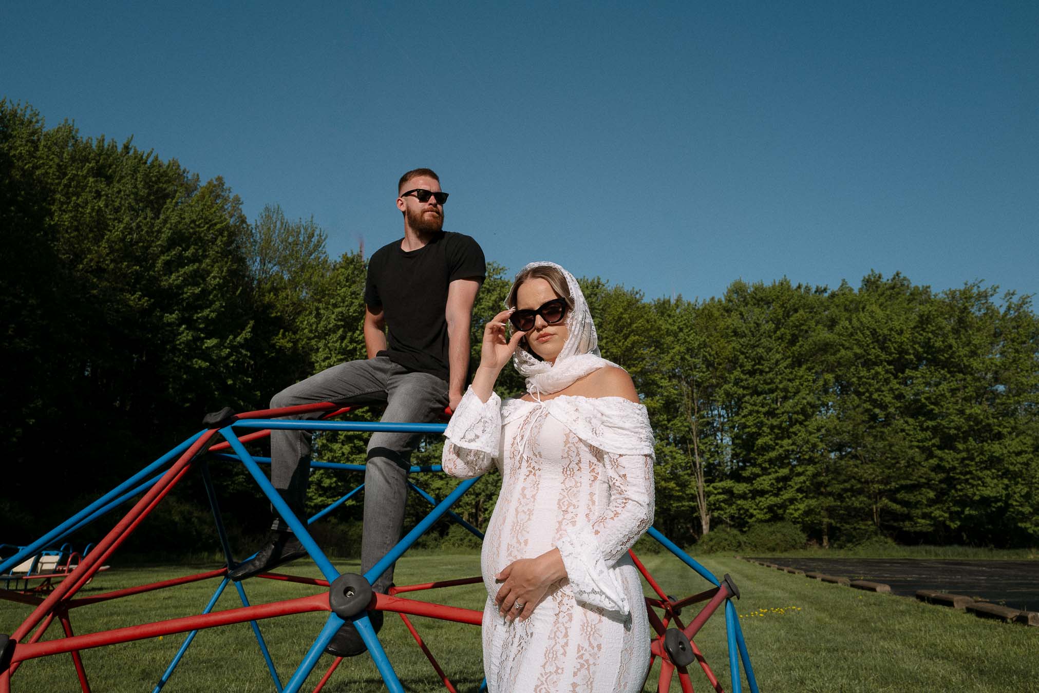 Two people on a geometric metal structure outdoors, with trees and a clear sky in the background.
