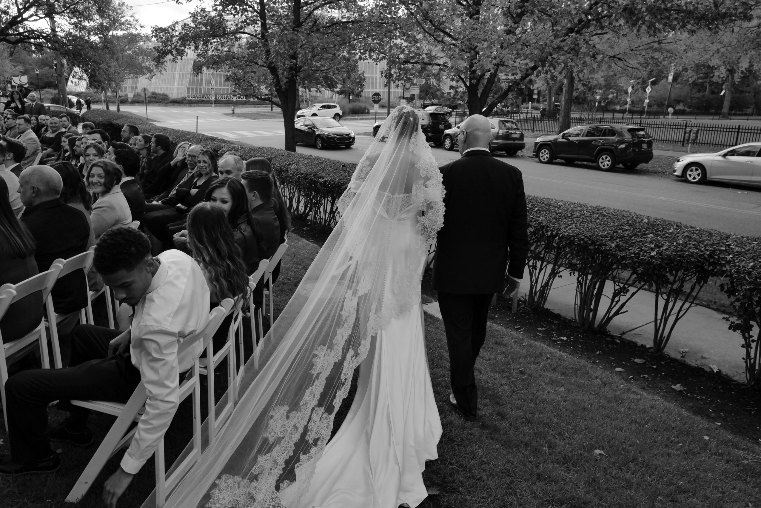 Bride in wedding dress and veil walking with a person in a suit outdoors, with seated people and cars in background.