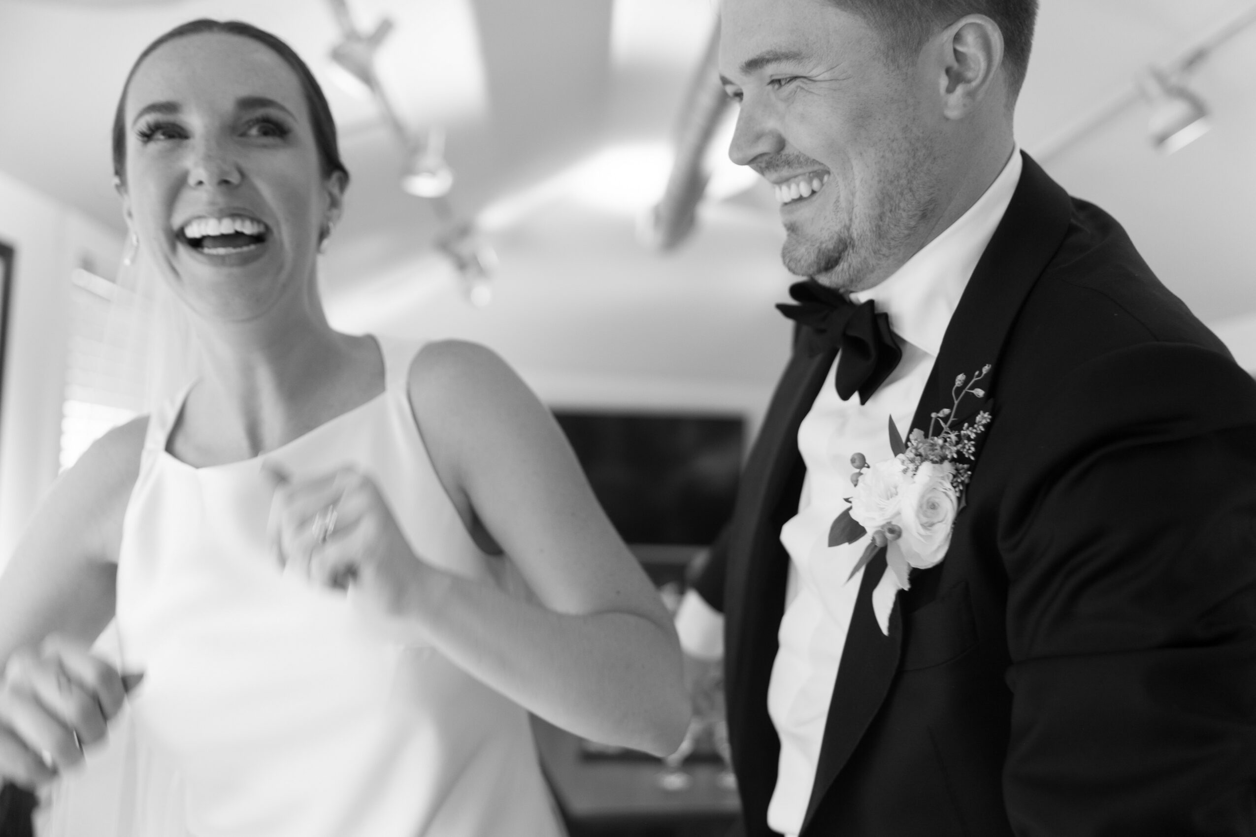 A smiling woman and a man in a tuxedo with a boutonniere share a joyful moment indoors.