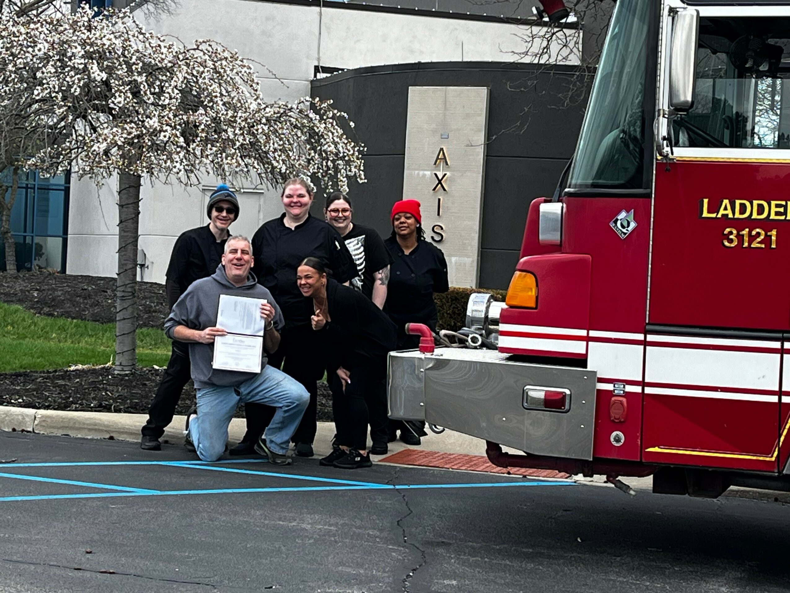 Group of six people posing next to a fire truck outside a building with a sign reading 'AXIS'. One person holds a certificate.
