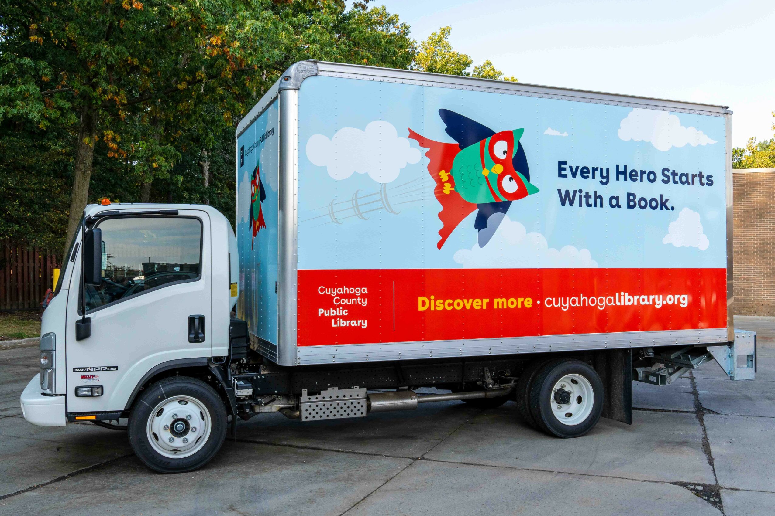 White delivery truck with a colorful fish mascot and a red banner promoting a library website, parked on a paved surface.