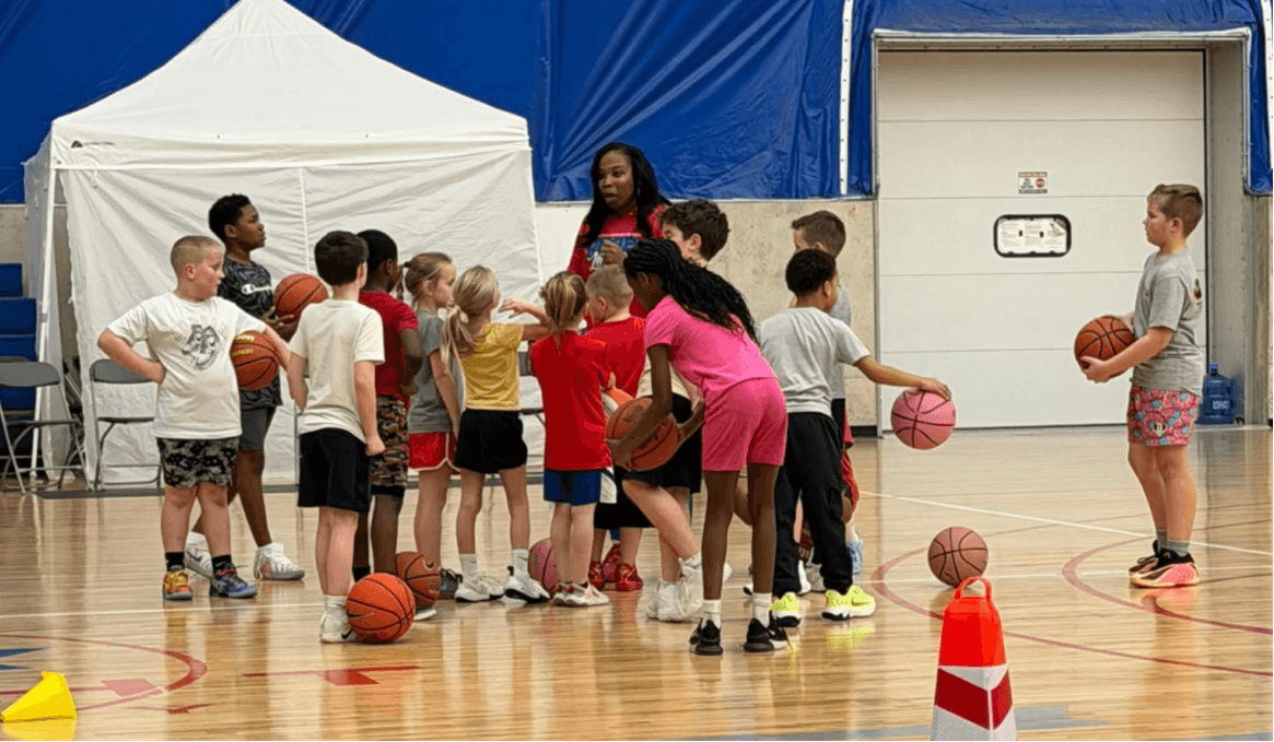 Group of children and two adults in a gymnasium, some holding basketballs, with a white tent and blue wall in background.