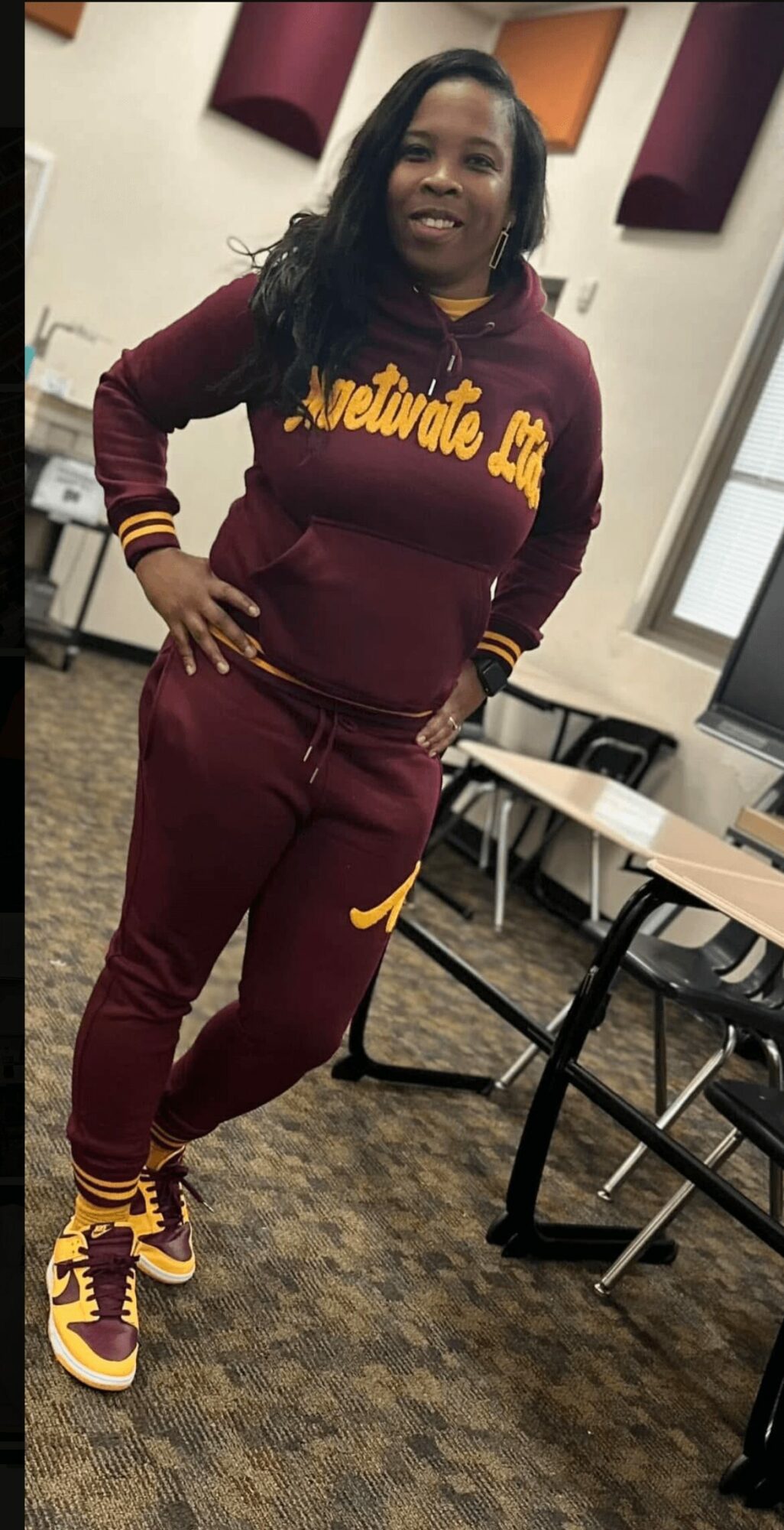 Woman in maroon and yellow athletic outfit standing in classroom with desks and chairs, smiling, hands on hips.