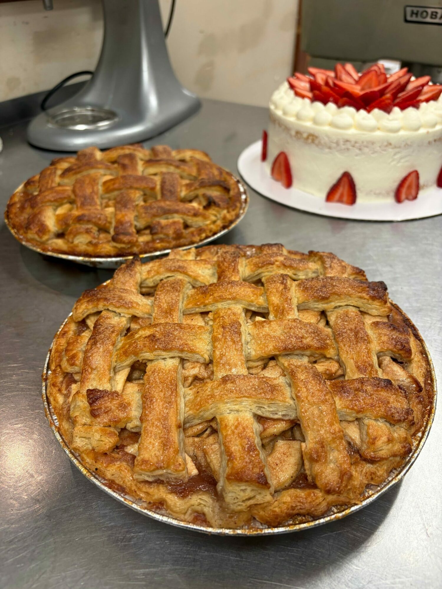 Two apple pies with lattice crusts on a table, one larger and one smaller, and a cake with strawberries in the background.