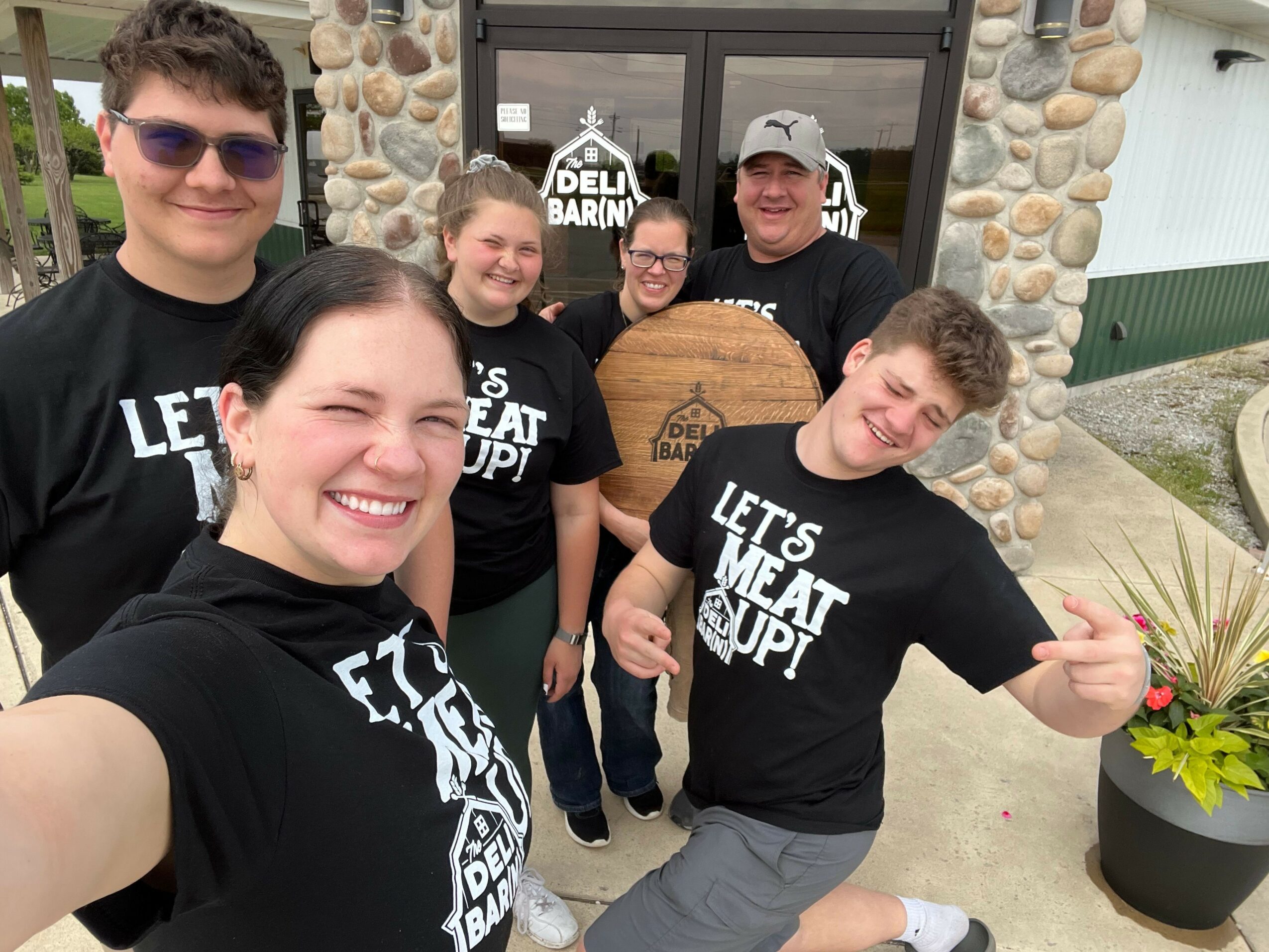 Four people smiling and taking a selfie outdoors, with a stone wall and window behind them.