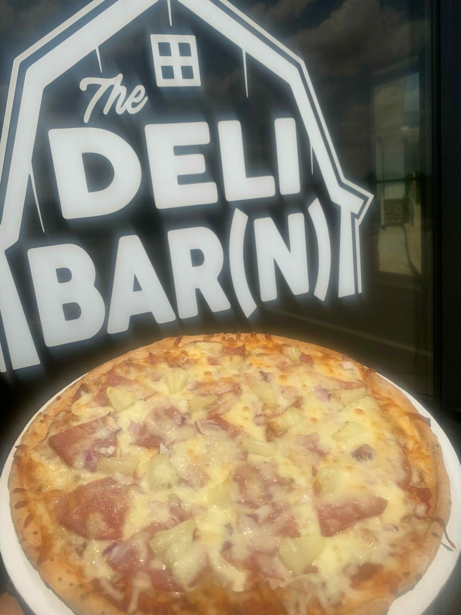 Pizza on a white plate with a cheese and meat topping, placed on a table near a window with a sign reading 'The DELI BAR' in the background.