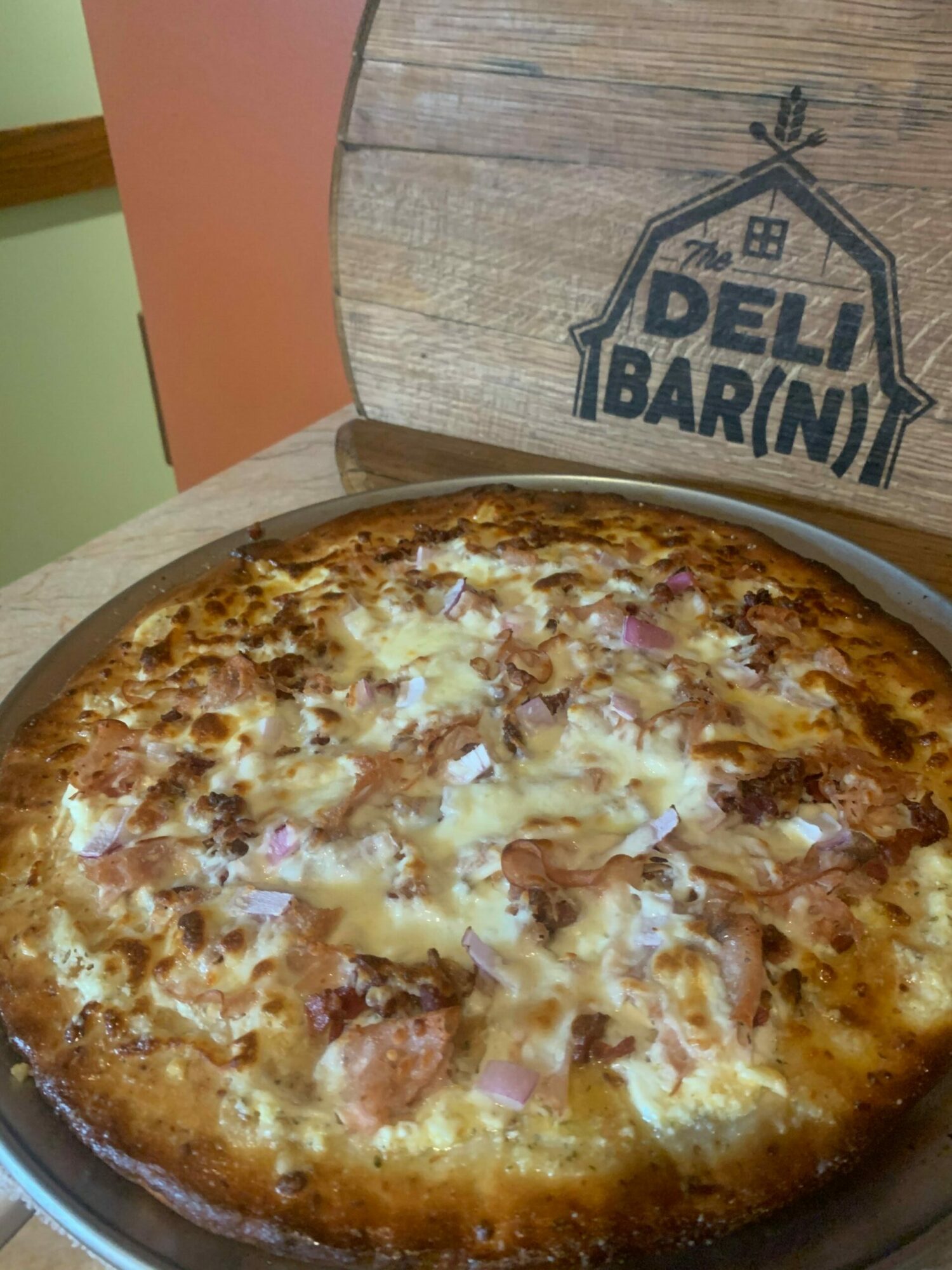 Pizza in a pan with cheese, ham, and onions, on a wooden table with a cardboard box nearby.