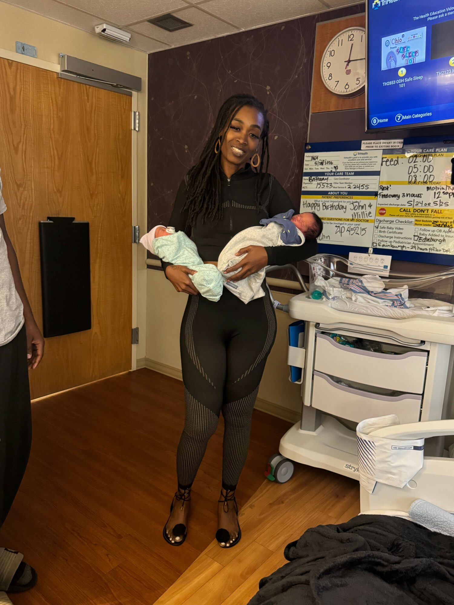 Woman holding two babies in a room with medical equipment and a computer monitor, standing on wooden flooring.