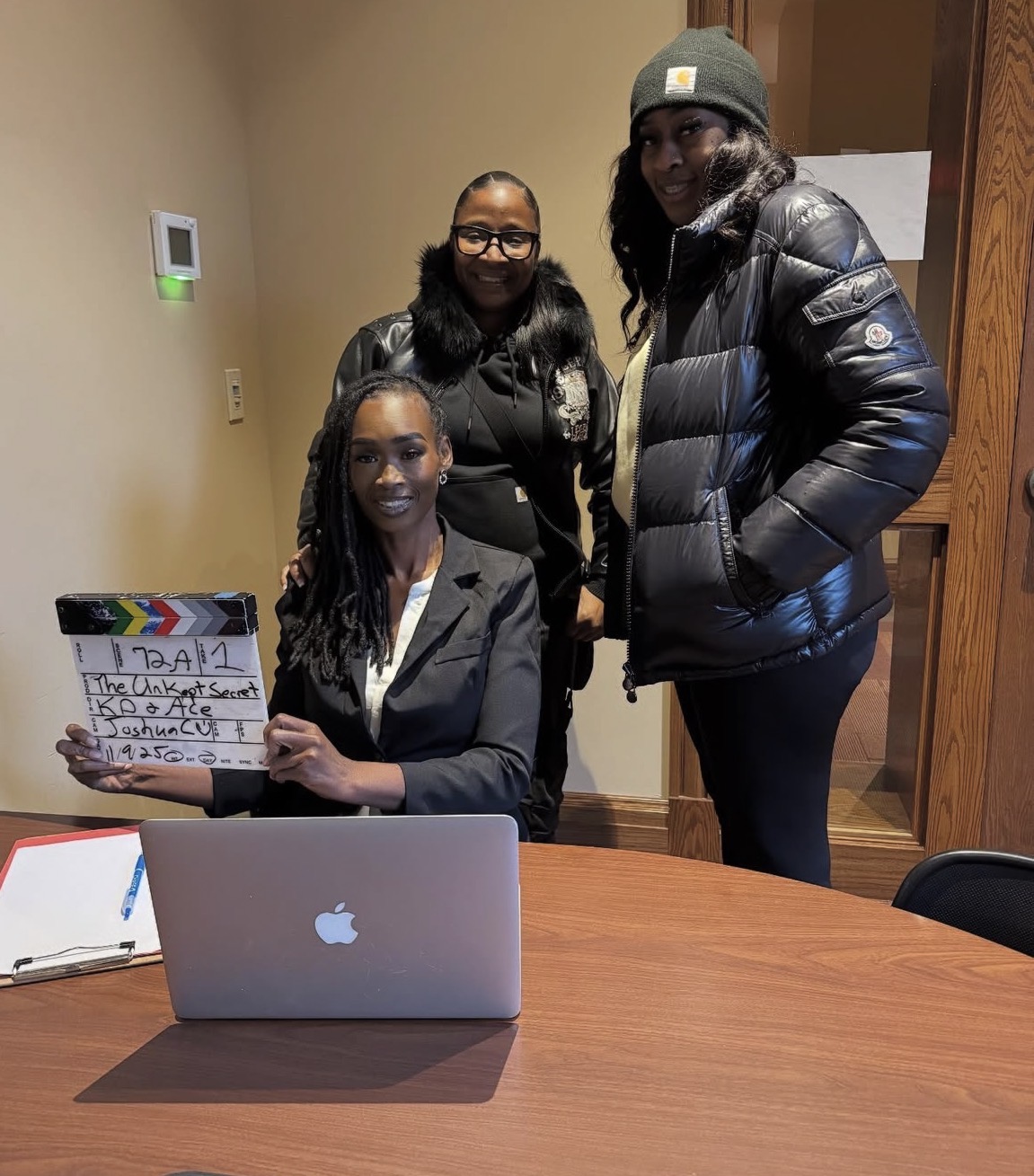 Three women in a room, one sitting at a table with a laptop and holding a clapperboard, two standing behind her, all smiling.