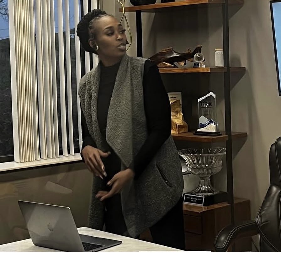 Woman standing near a bookshelf with a laptop on a table in front of her.