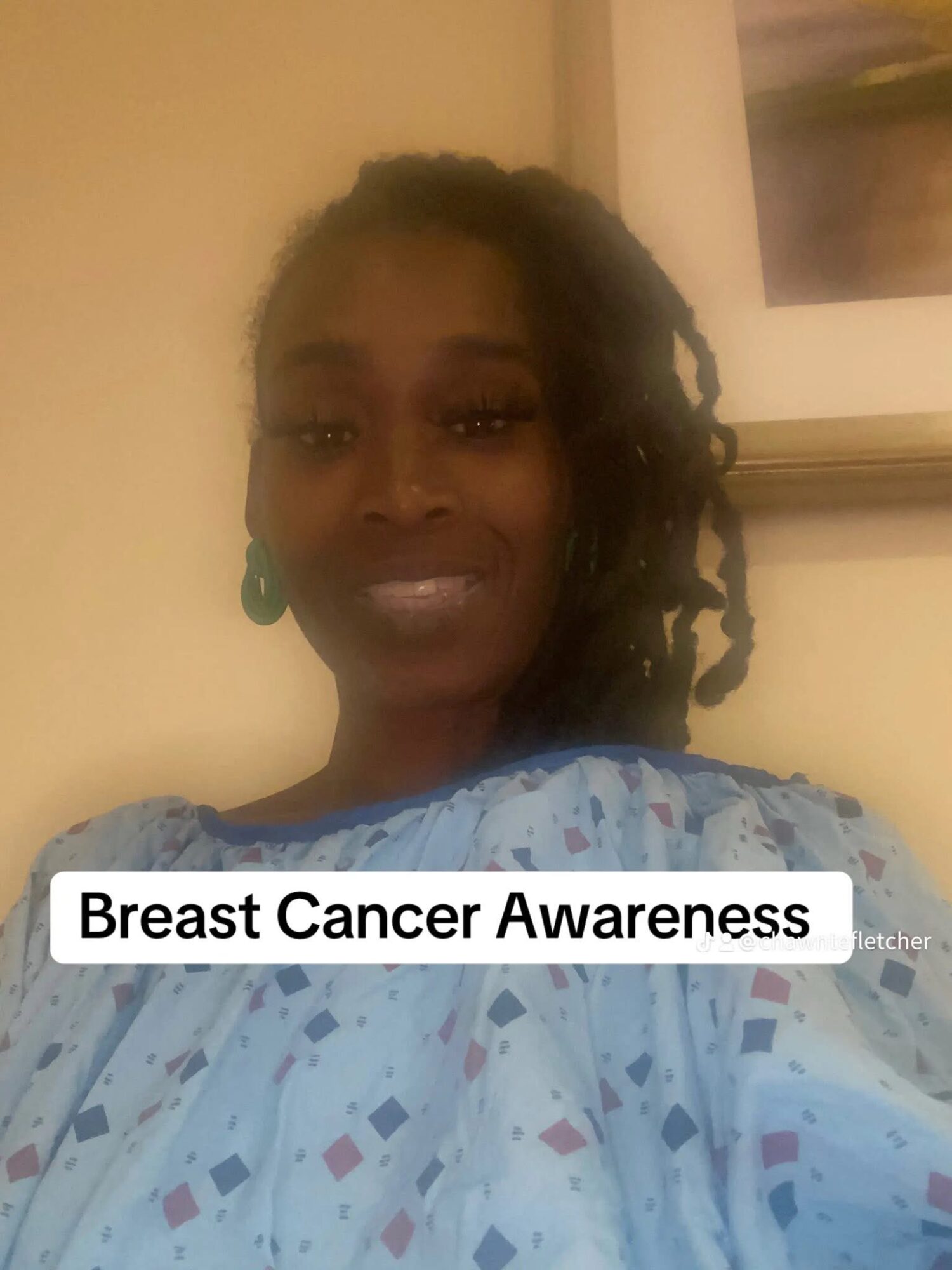 Woman in hospital gown smiling, promoting breast cancer awareness, with a framed picture in the background.
