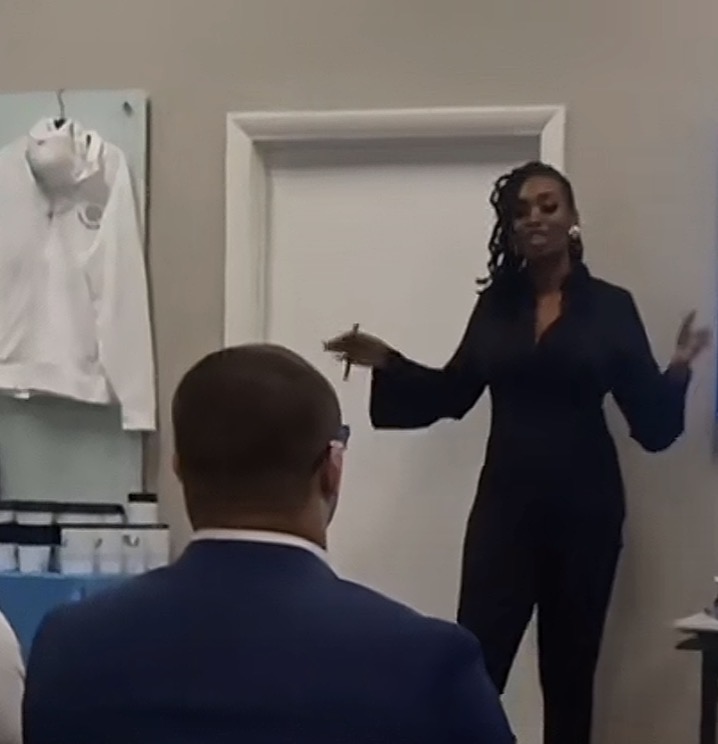Woman standing and speaking to a seated man in a suit, in a room with a white door and clothing on the wall.