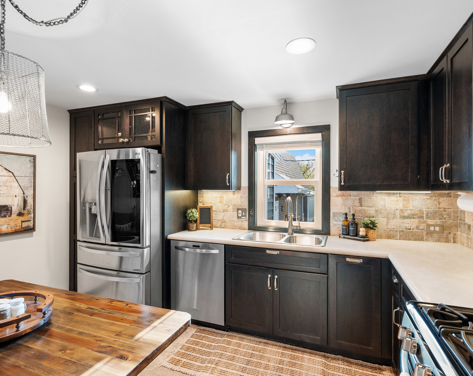 Kitchen with dark cabinets, stainless steel refrigerator, window above sink, beige backsplash, and wooden table in foreground.
