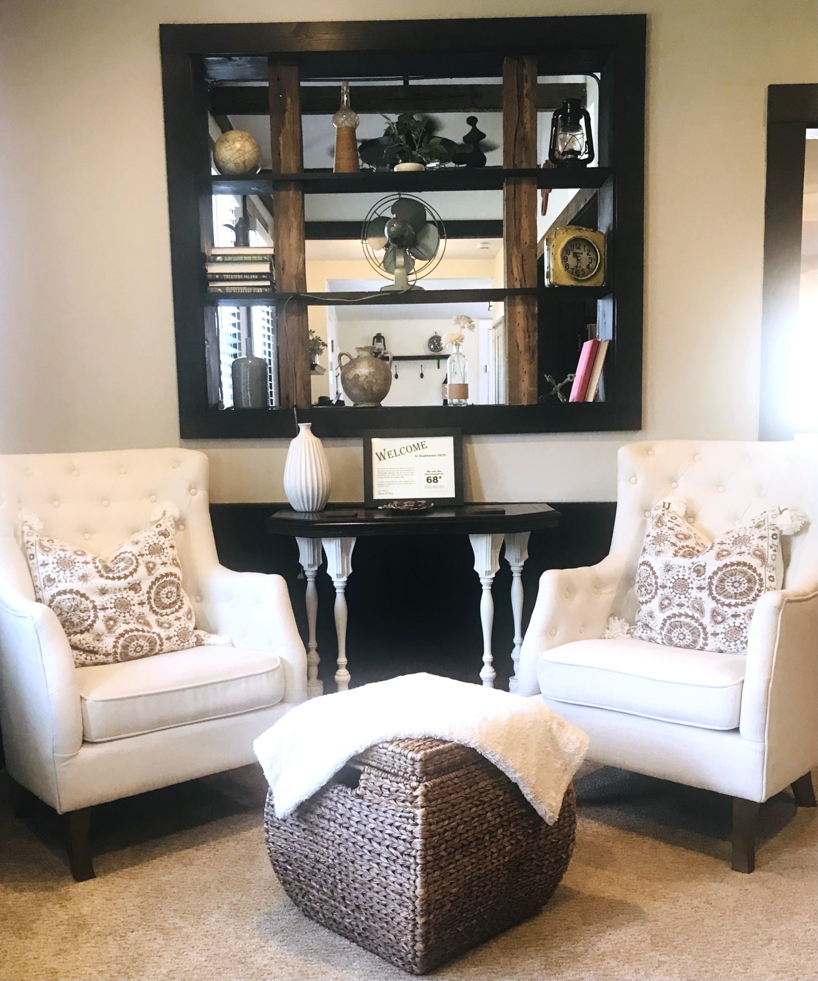Two white armchairs with patterned pillows face each other, with a woven ottoman in between, in a cozy room with a black shelf and table behind.