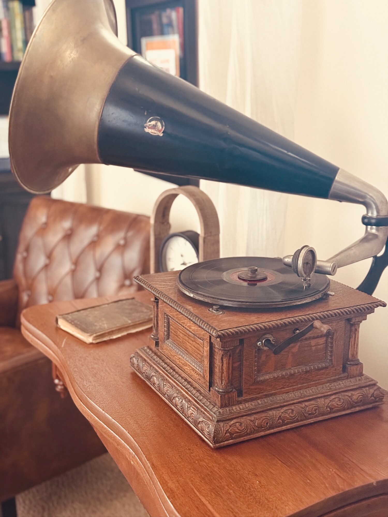 Vintage gramophone with a large black horn on a wooden table, with a leather chair and books in the background.