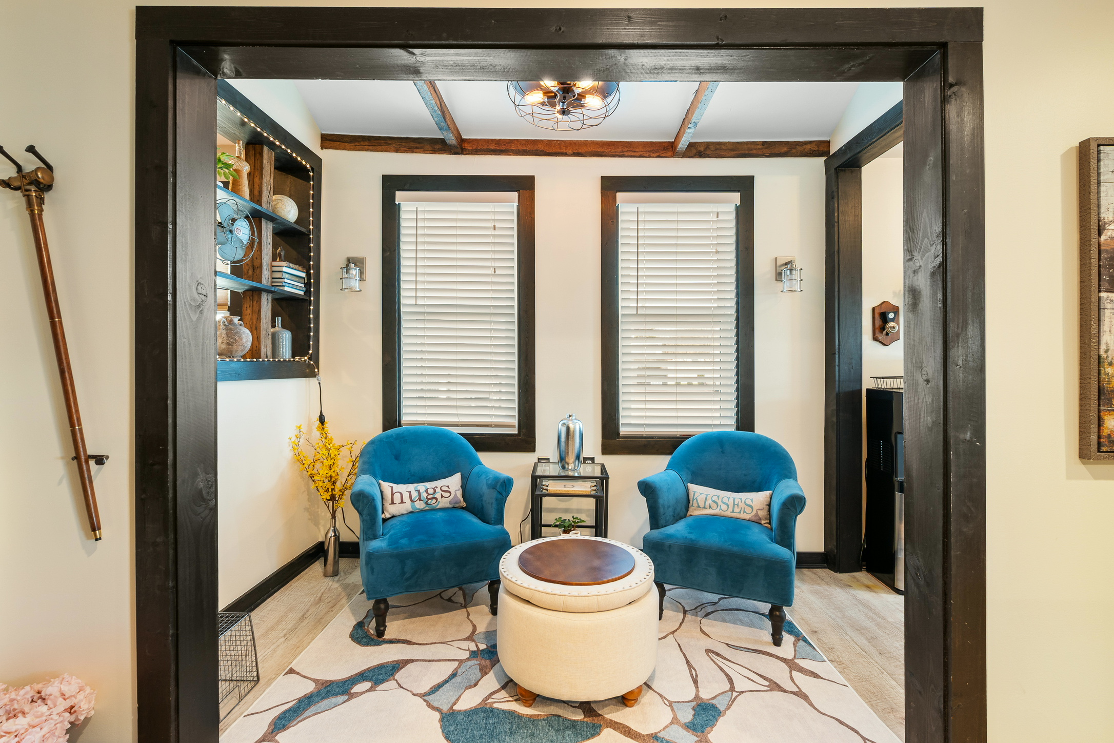 Living room with two blue armchairs, a white ottoman, and two windows with blinds, viewed through a doorway.