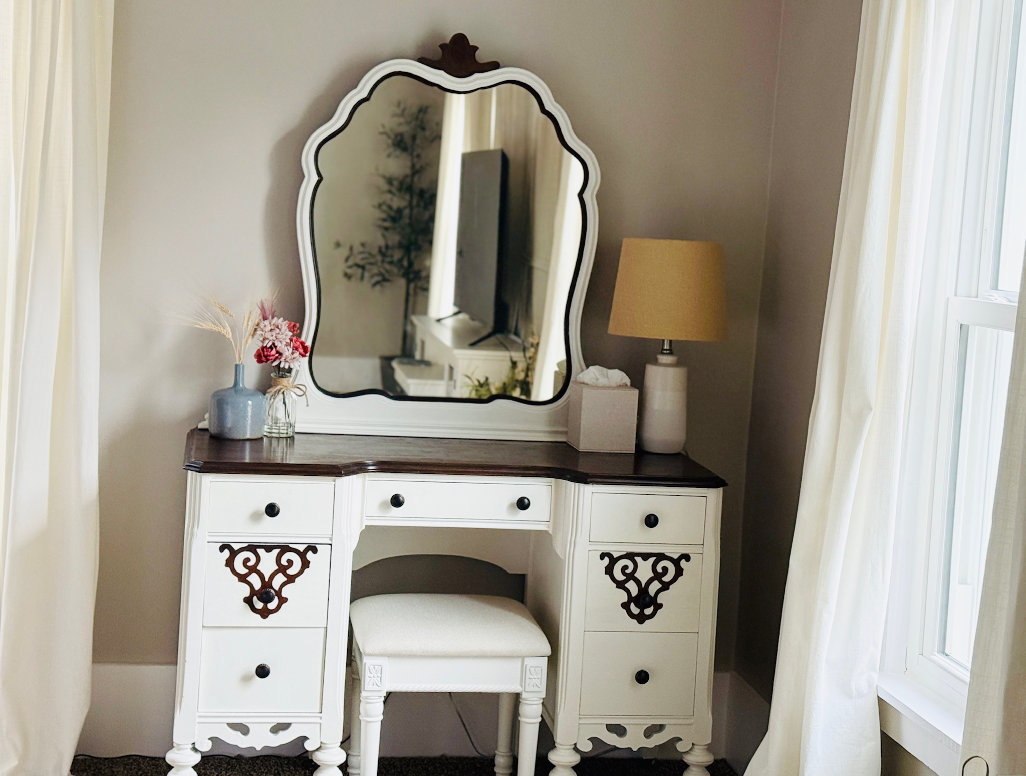 White vanity table with a mirror, decorated with a lamp, flowers, and a small stool, in a bright room.