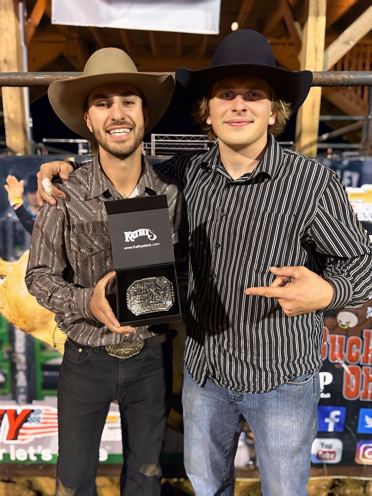 Two men wearing cowboy hats and striped shirts stand together, one holding a box with a shiny object inside.