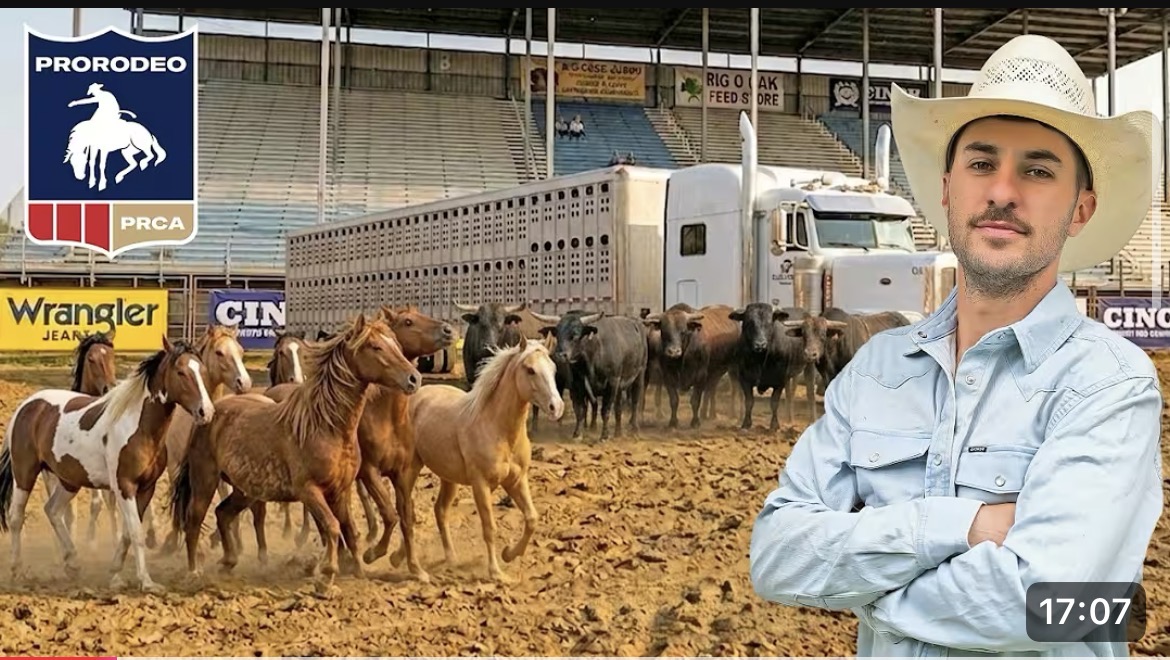 Man in cowboy hat standing with arms crossed in front of a cattle herd at an indoor rodeo arena.