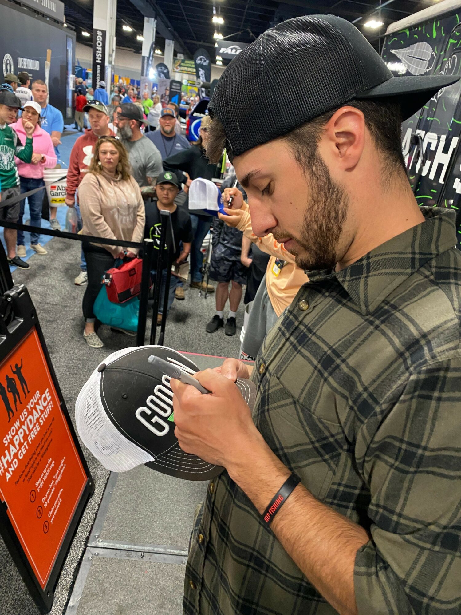 Man with a beard and cap holding a white and black object, standing in a busy indoor setting with people and shopping carts.