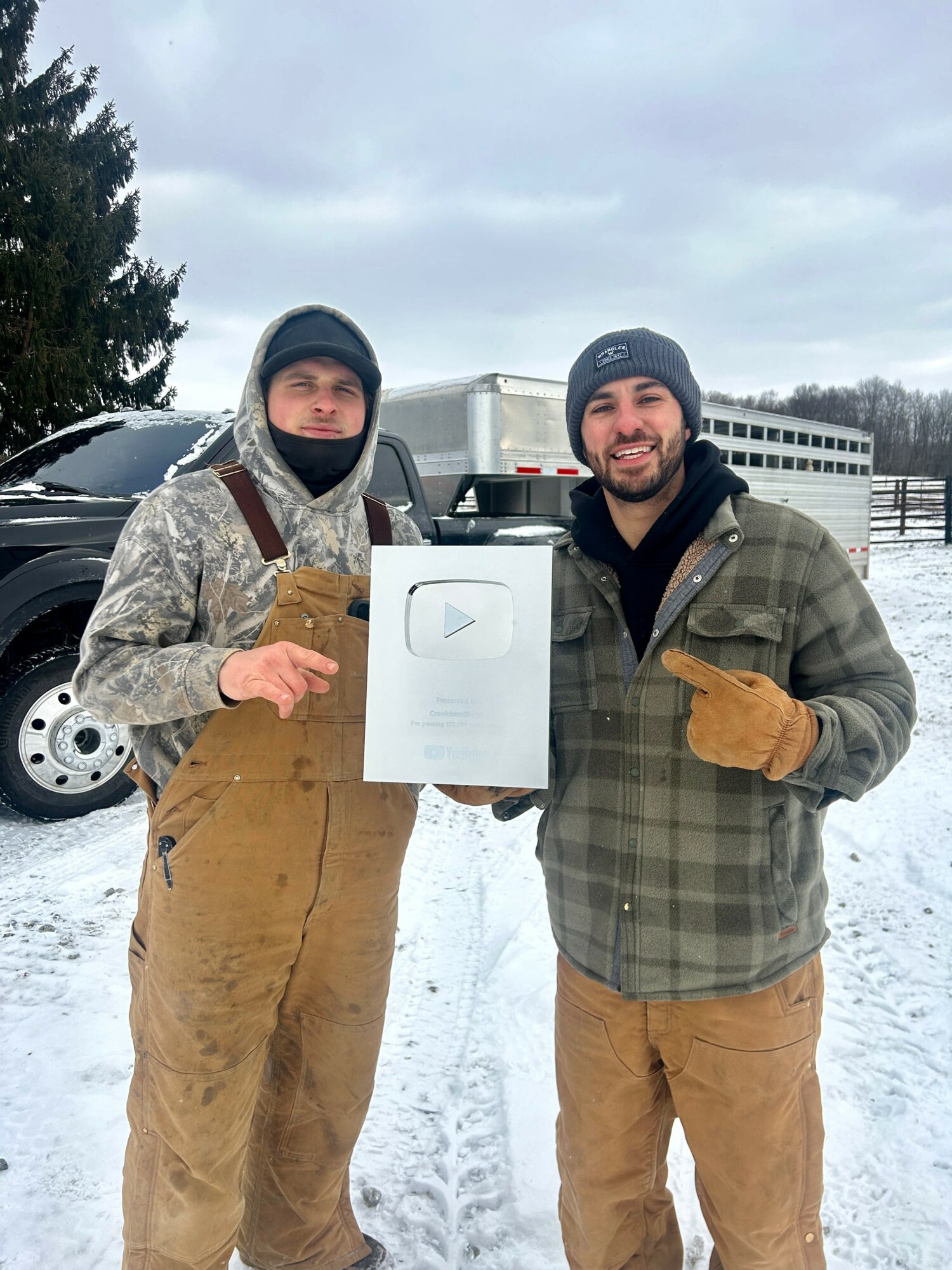 Two men outdoors in winter, one holding a framed certificate, both smiling, wearing warm clothing, snow on ground, buildings in background.