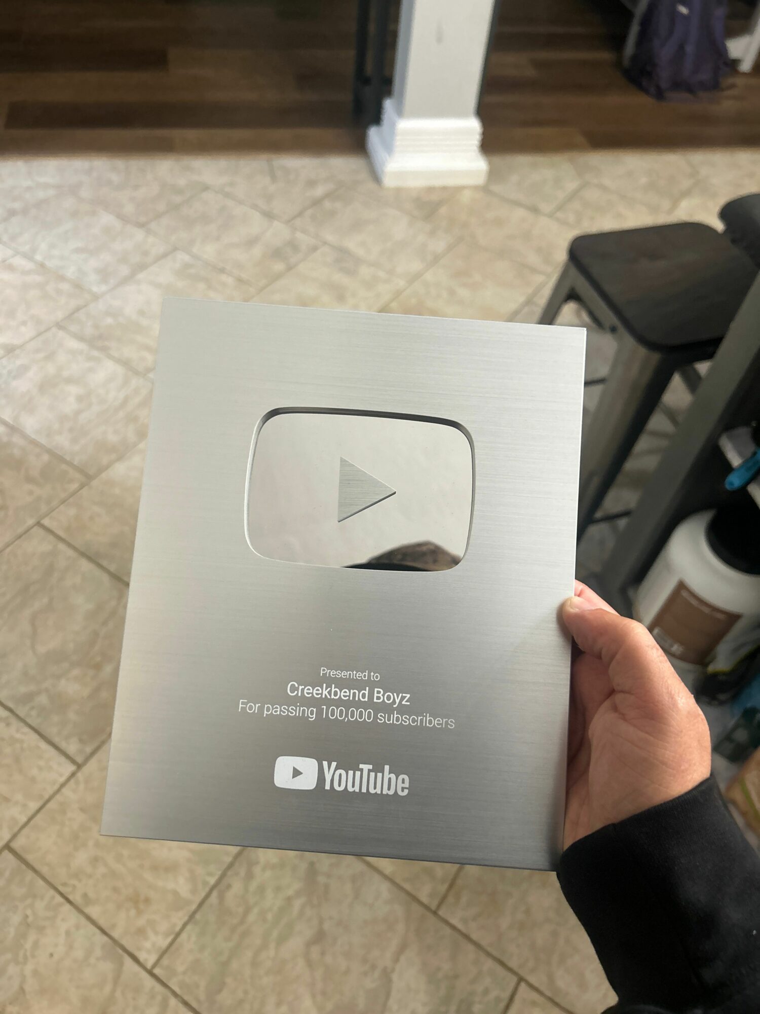 Person holding a YouTube Creator Award plaque in a room with tiled floor and shelves.