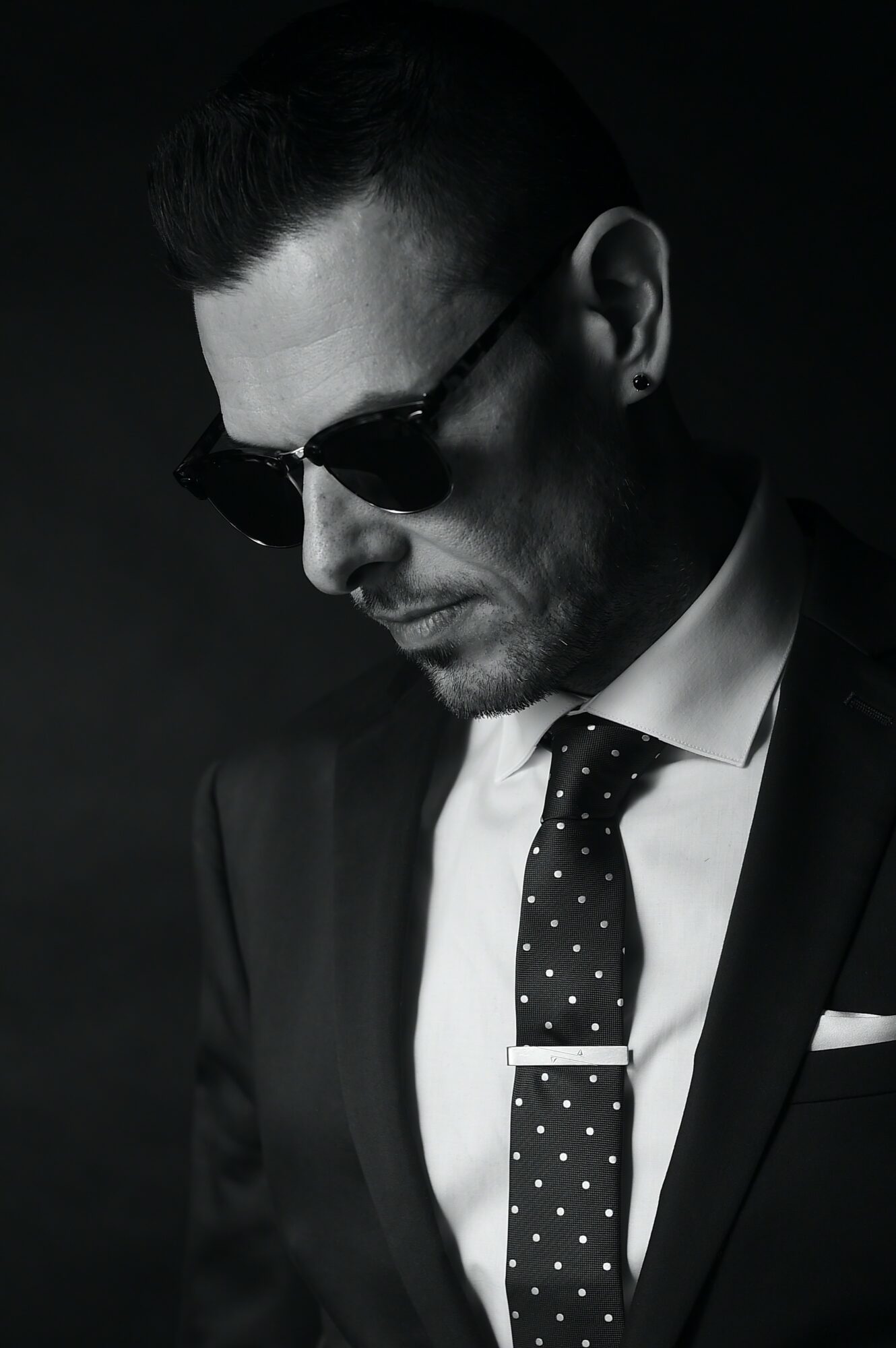 Man in suit wearing sunglasses, looking down, with dark background, black and white photo.