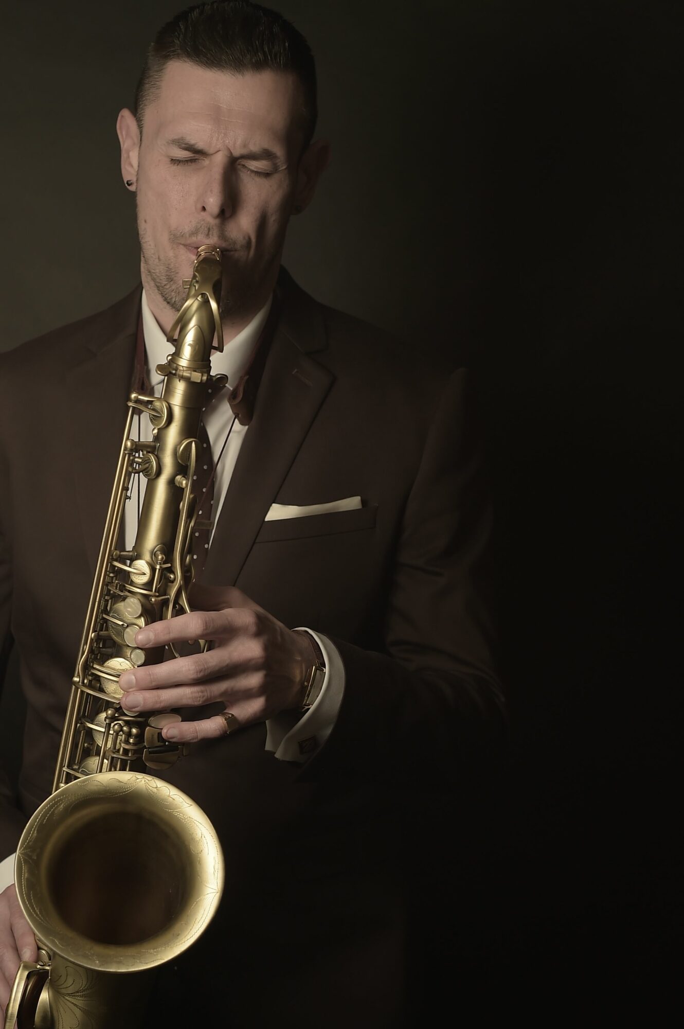 Person in dark suit playing a saxophone against a dark background, eyes closed, focused expression.