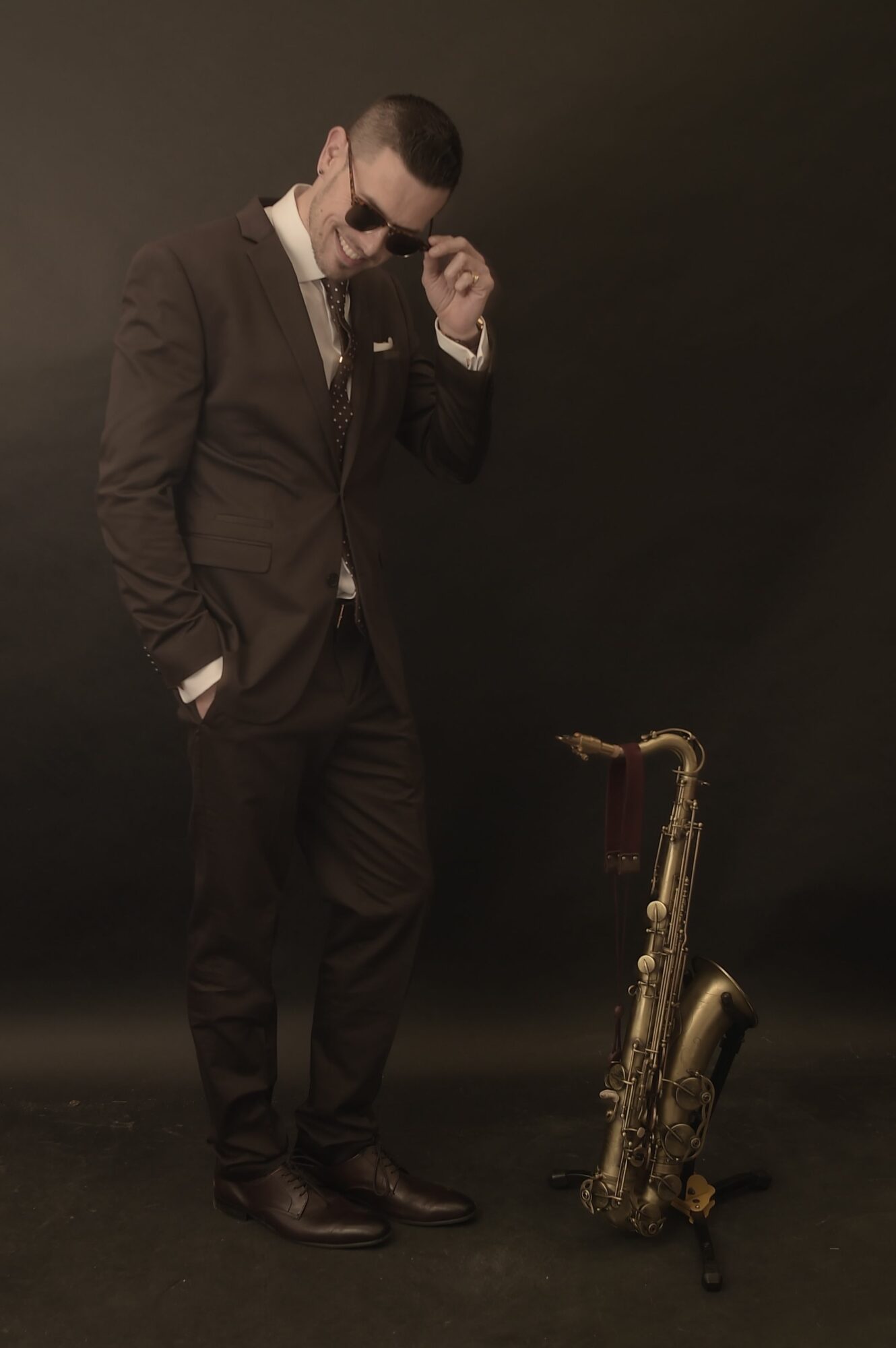Man in a suit holding a microphone, standing next to a saxophone on the floor, against a dark background.