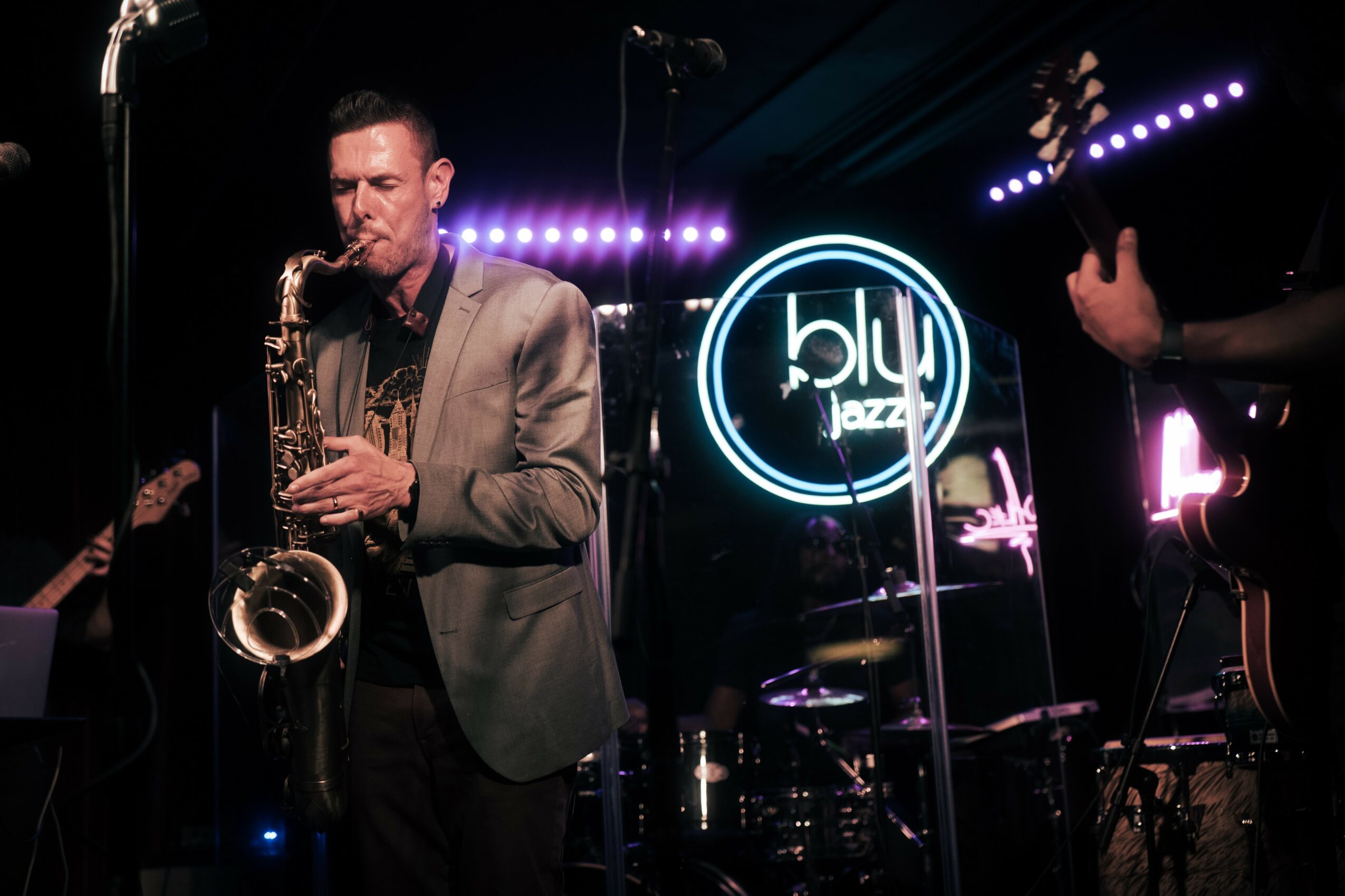 A man playing saxophone on stage with a blue jazz sign in the background, stage lights, and musical instruments.