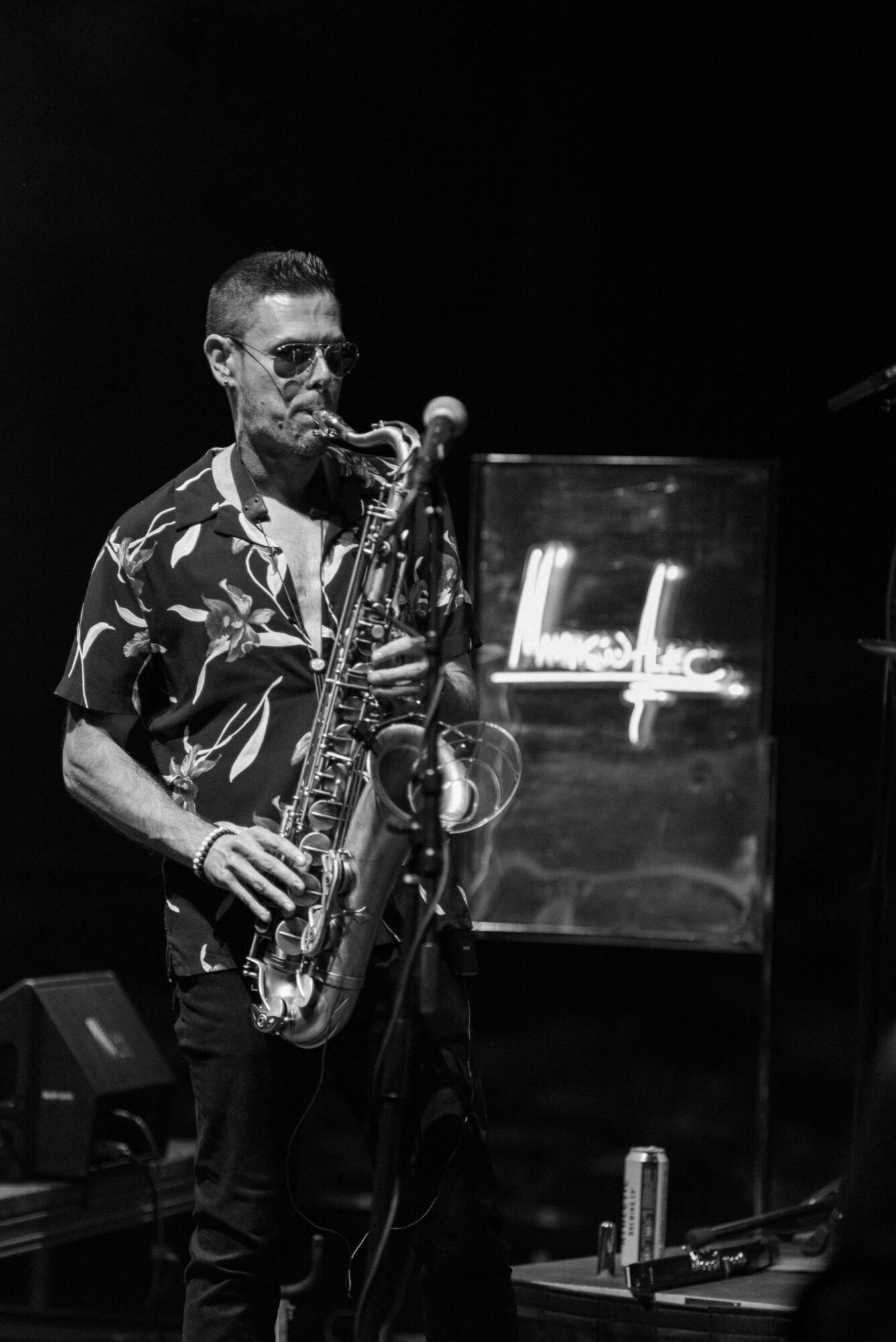 A man playing a saxophone on stage with a neon sign in the background, wearing sunglasses and a patterned shirt.