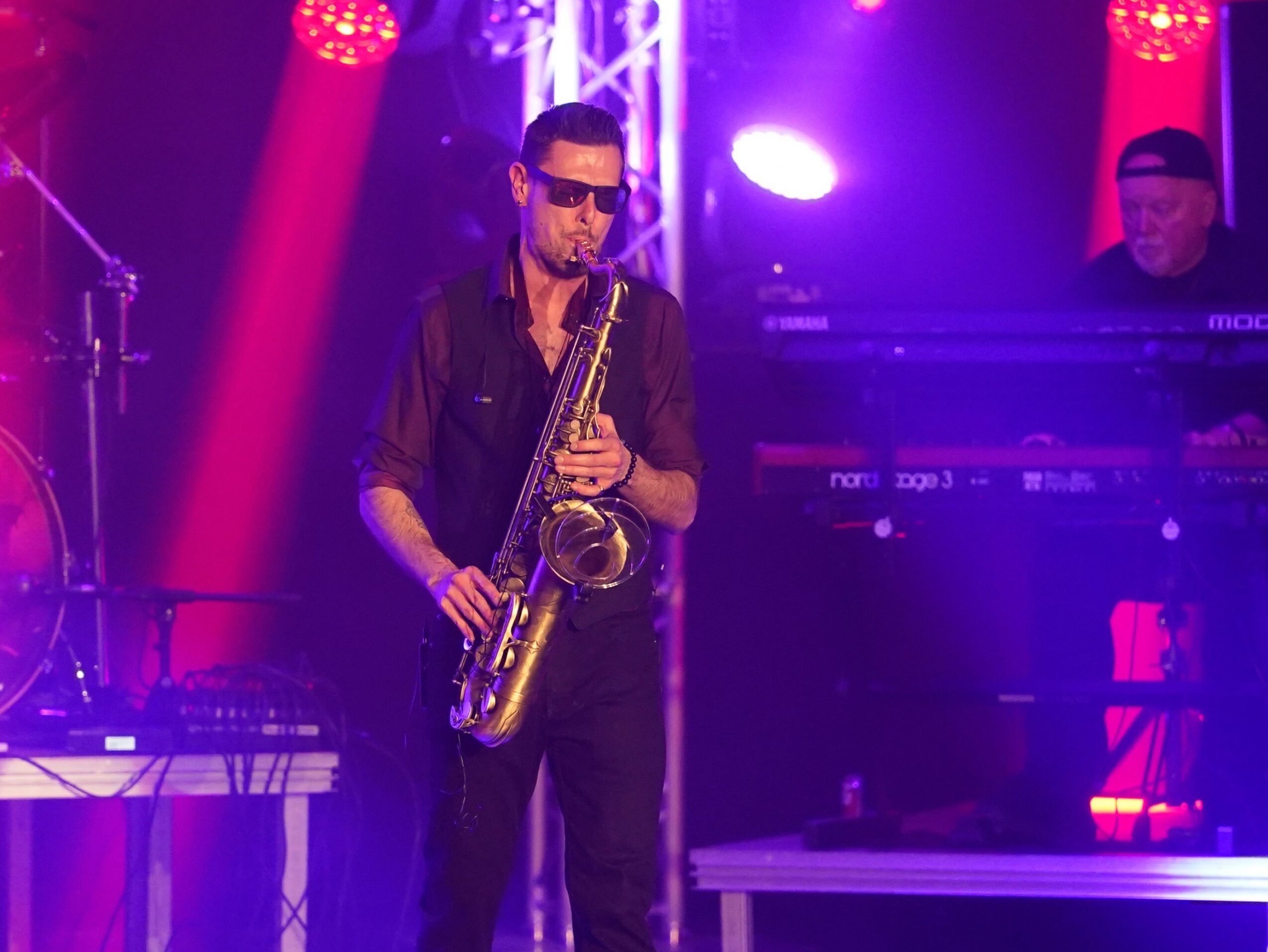 Musician playing saxophone on stage with purple and red lighting, keyboard player in background, dark clothing, sunglasses.