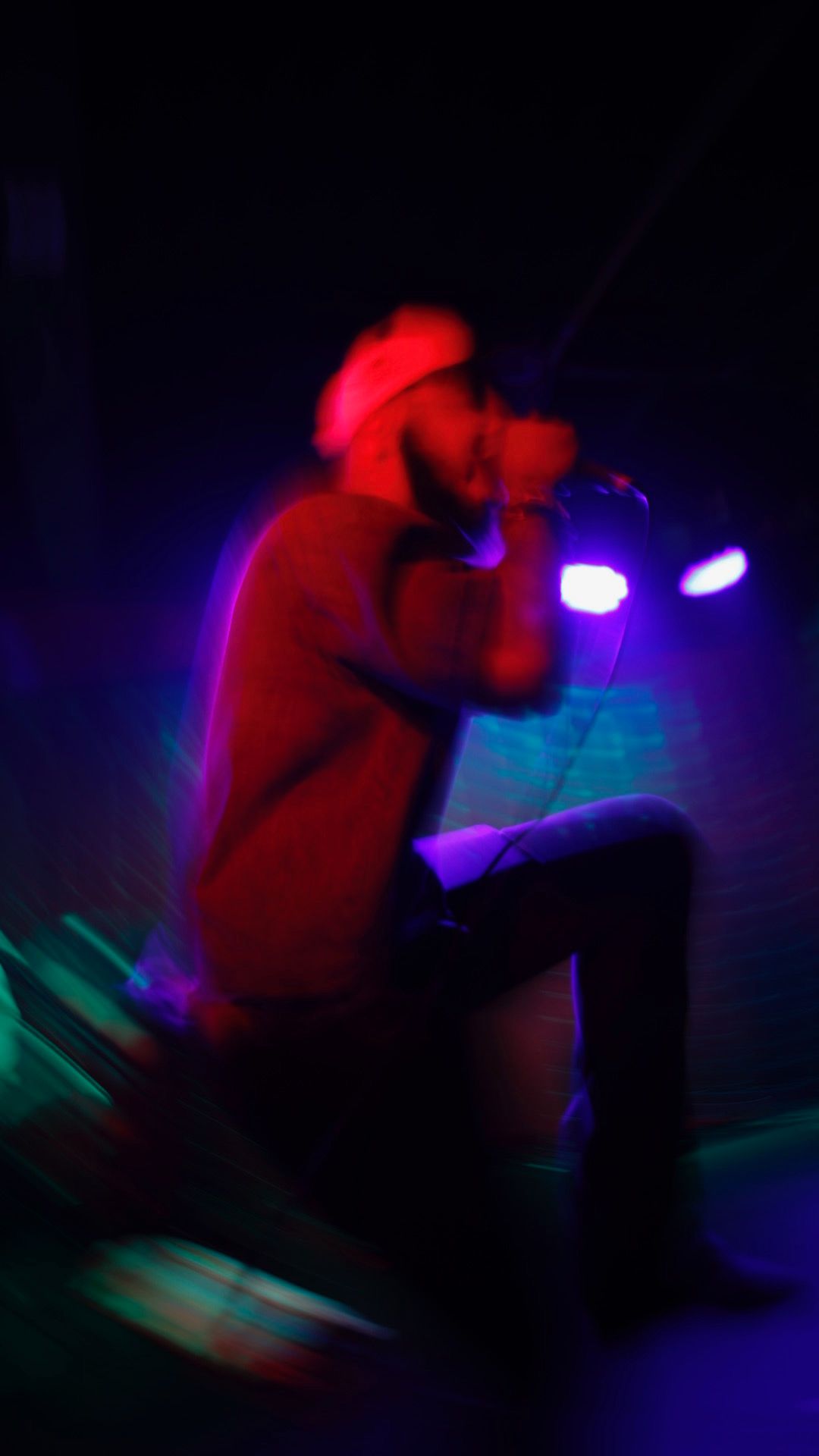 Person crouching in a dark room with colorful lights, wearing a red hoodie and cap.