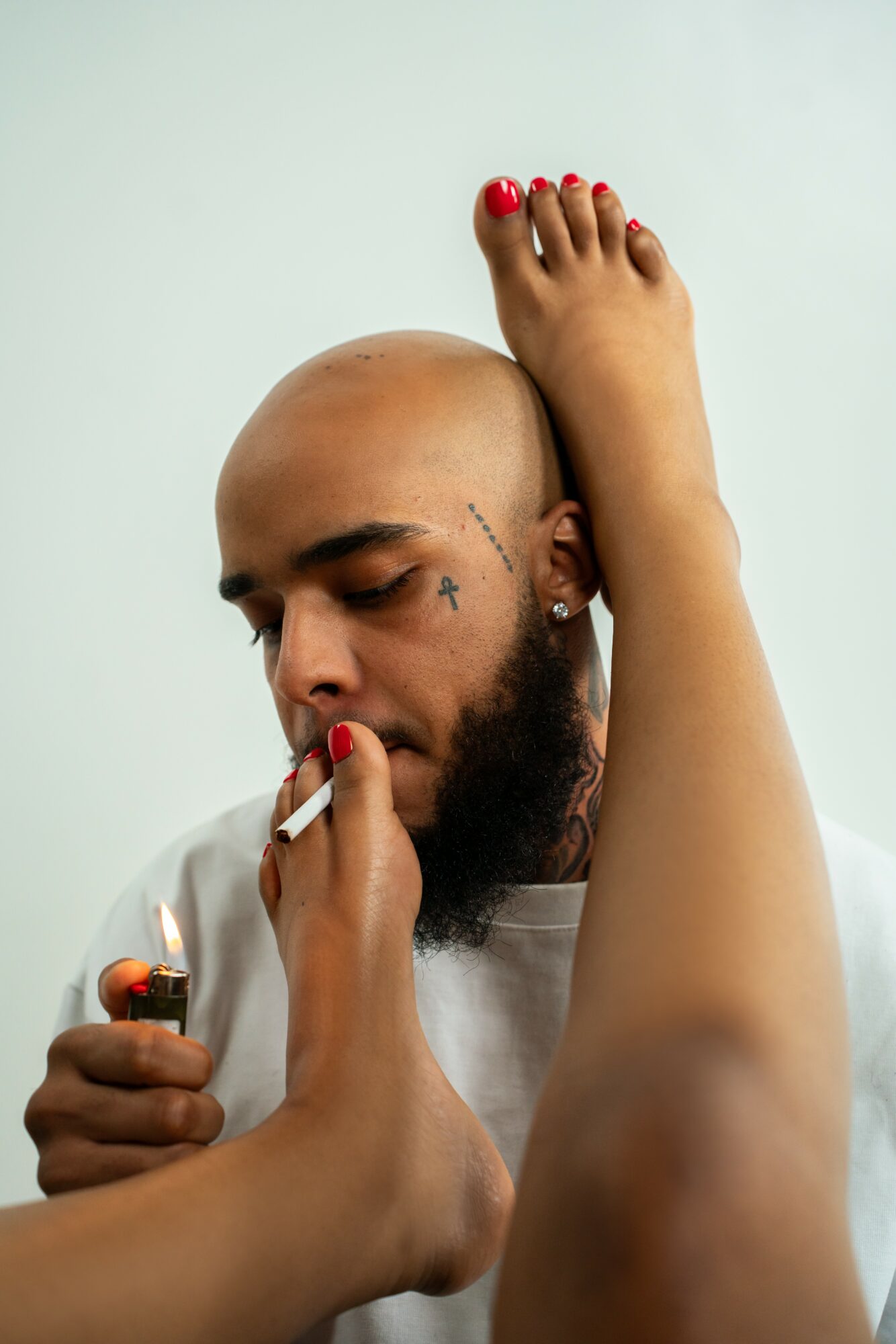 Man with a beard and tattoo on face, eyes closed, receiving a pedicure, foot with red nail polish near face.