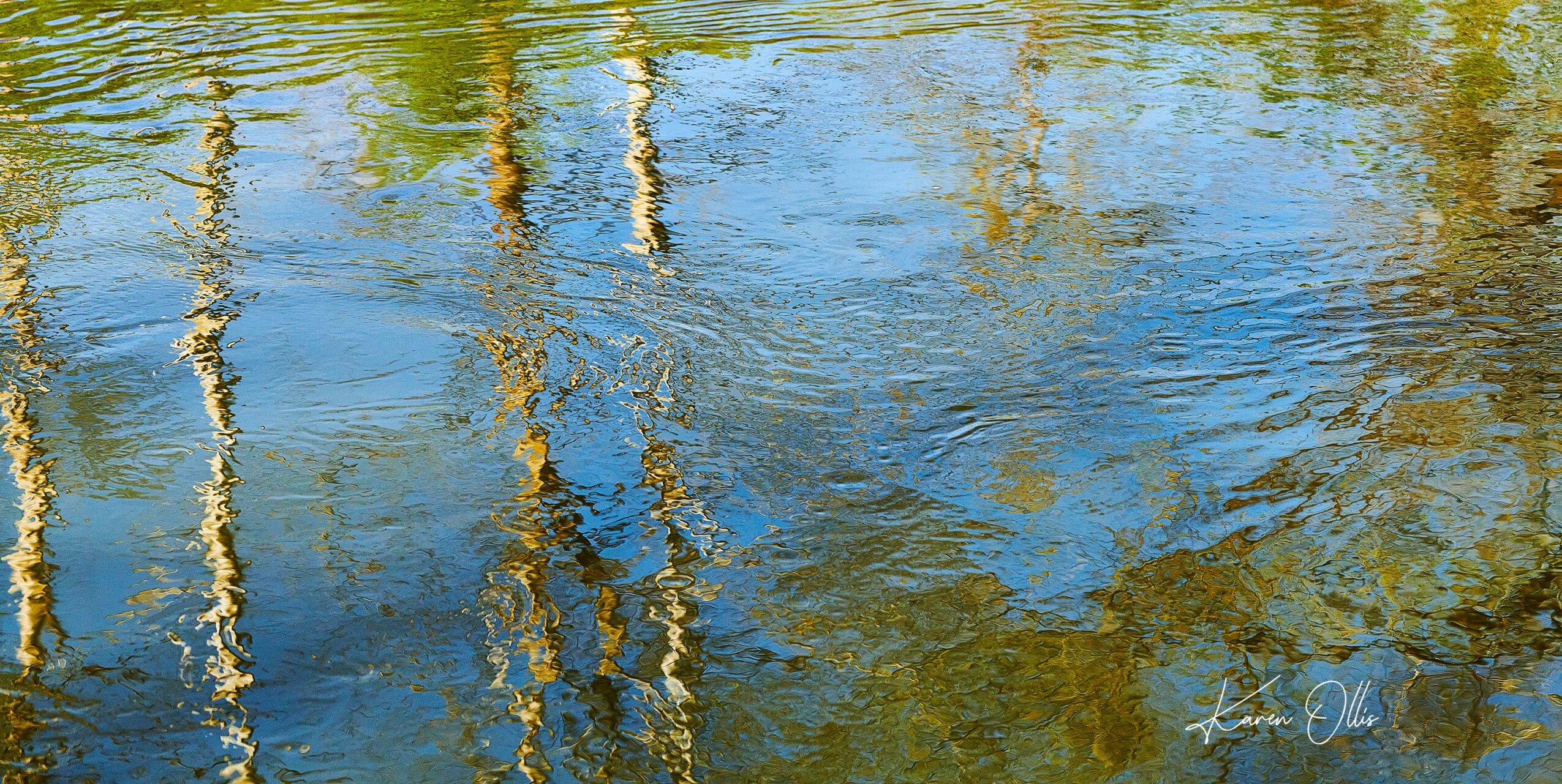 Reflections of trees and sky on a rippling water surface, with blue, green, and yellow colors.