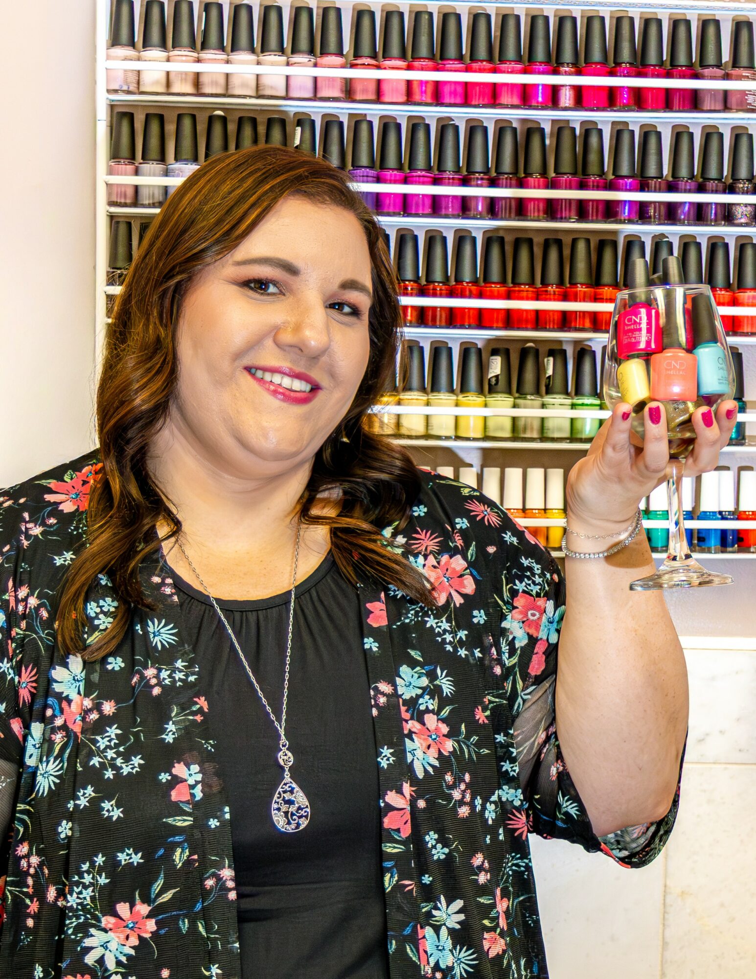 Woman with long brown hair holding nail polish bottles in front of a wall of nail polish colors, smiling, wearing a floral jacket and necklace.