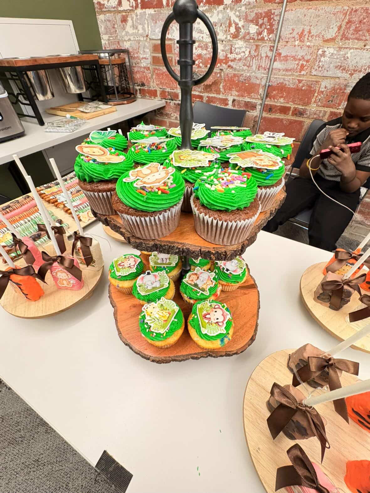 Cupcakes with green frosting and holiday-themed decorations on a tiered stand, surrounded by other desserts on plates, in a room with brick wall.
