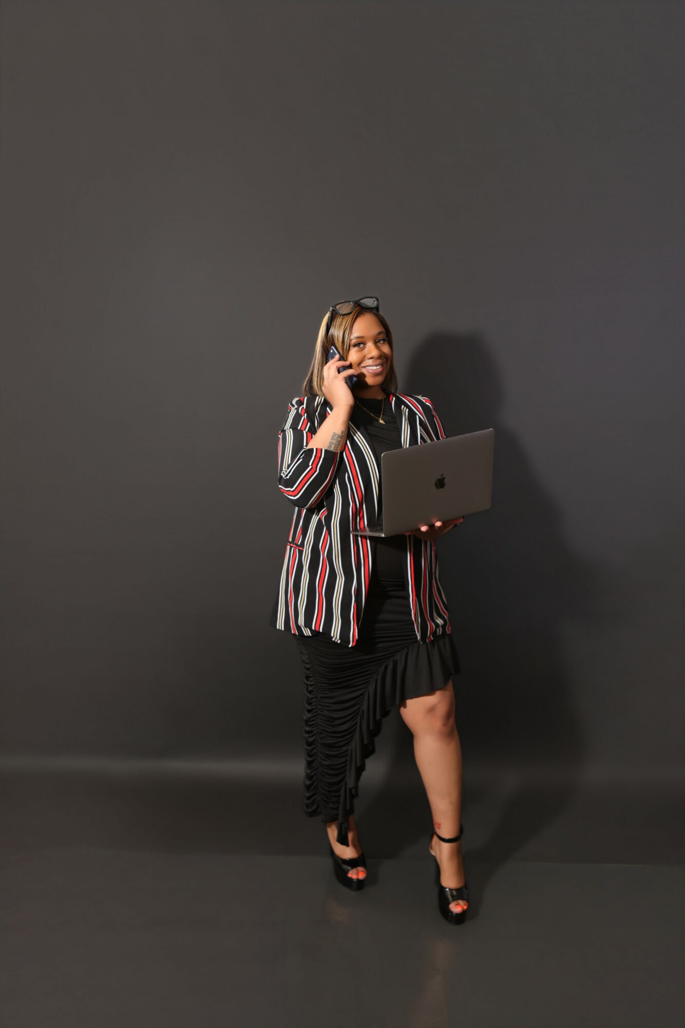 Woman standing with a laptop, talking on a phone, wearing a striped blazer and black dress, against a dark background.