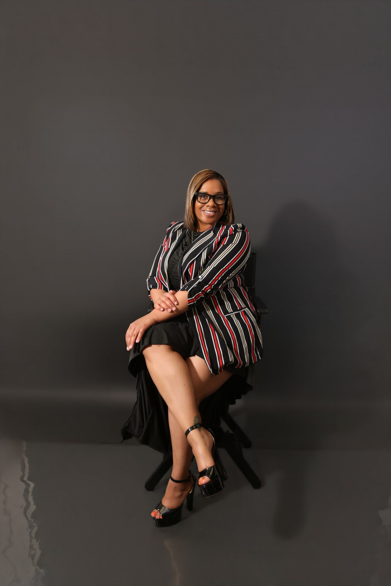 Woman sitting on a chair with crossed legs, wearing glasses and a striped blazer, against a plain dark background.