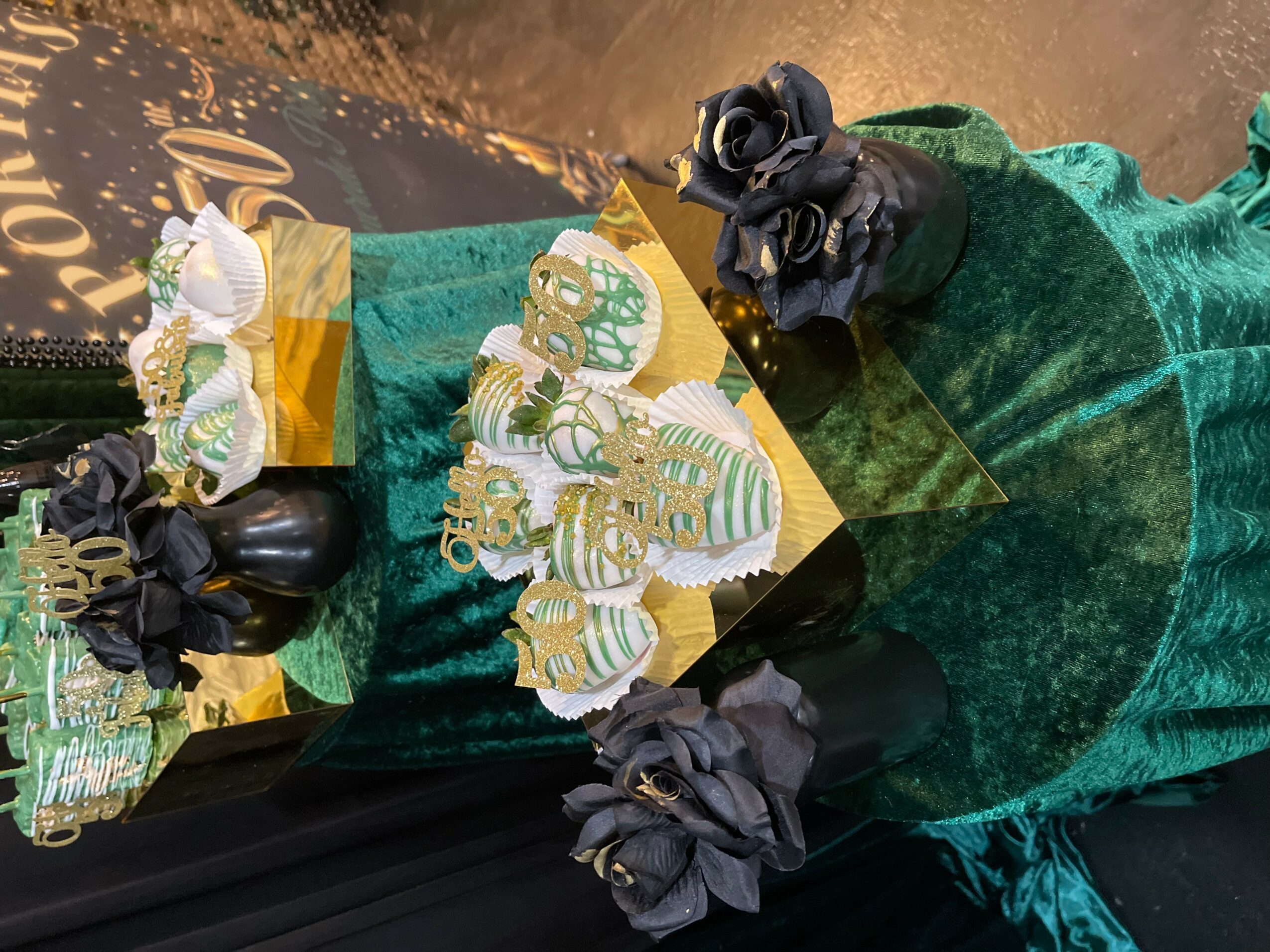 Decorative cupcakes with gold and white frosting, black roses, and gold accents on a green velvet cloth.
